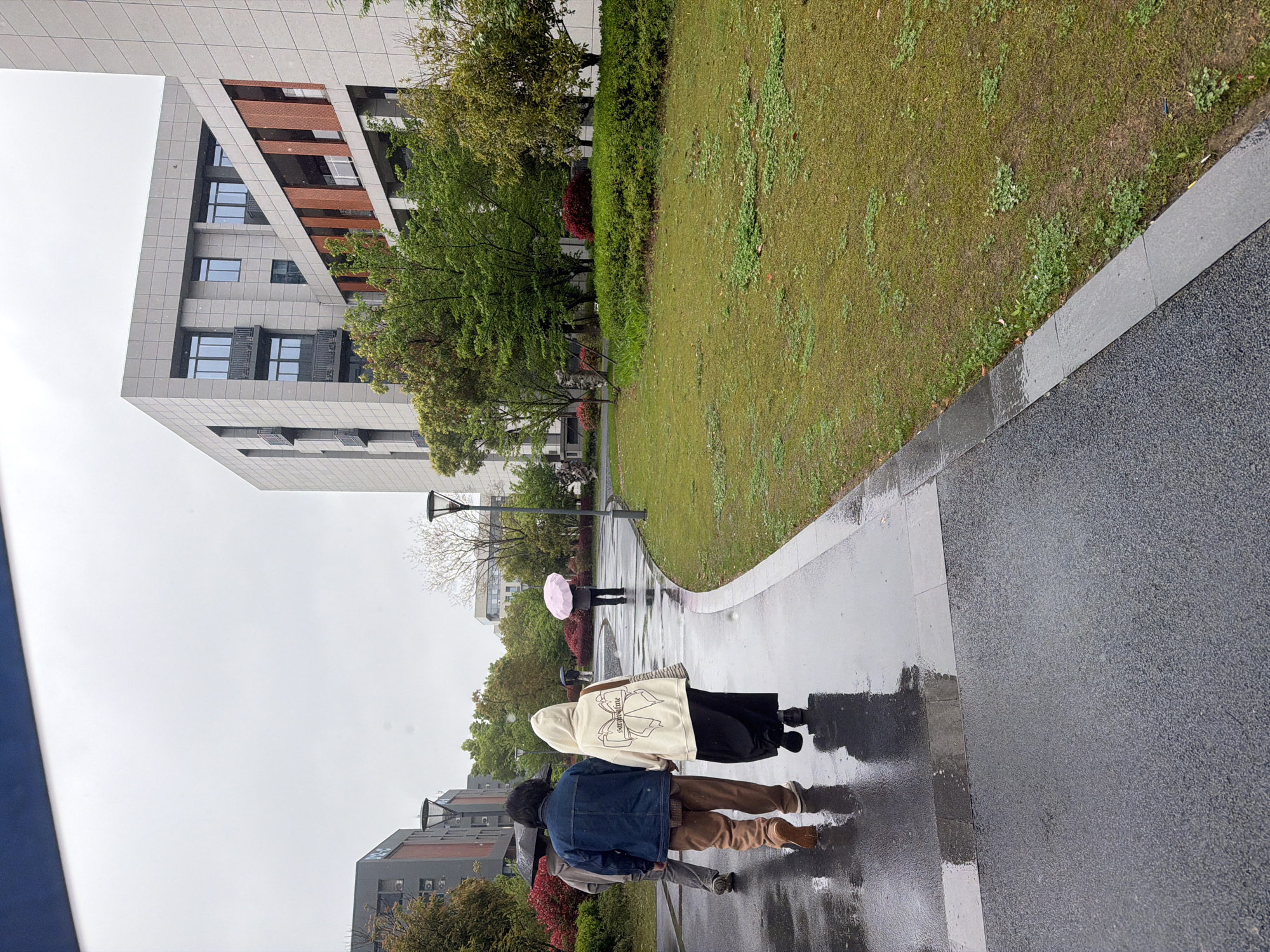 The image depicts a wet paved walkway in an urban environment, likely after or during rainfall. Two individuals walk along the walkway in the foreground. The first person wears a blue denim jacket and brown pants. The second person is wearing a white hooded sweatshirt and a black skirt. Further in the distance, a third person walks under a pink umbrella. A building is located on the left side of the image. The walkway is lined by a grassy area with trees and bushes. Another building is located behind the trees. The location is Jiangning District, China.