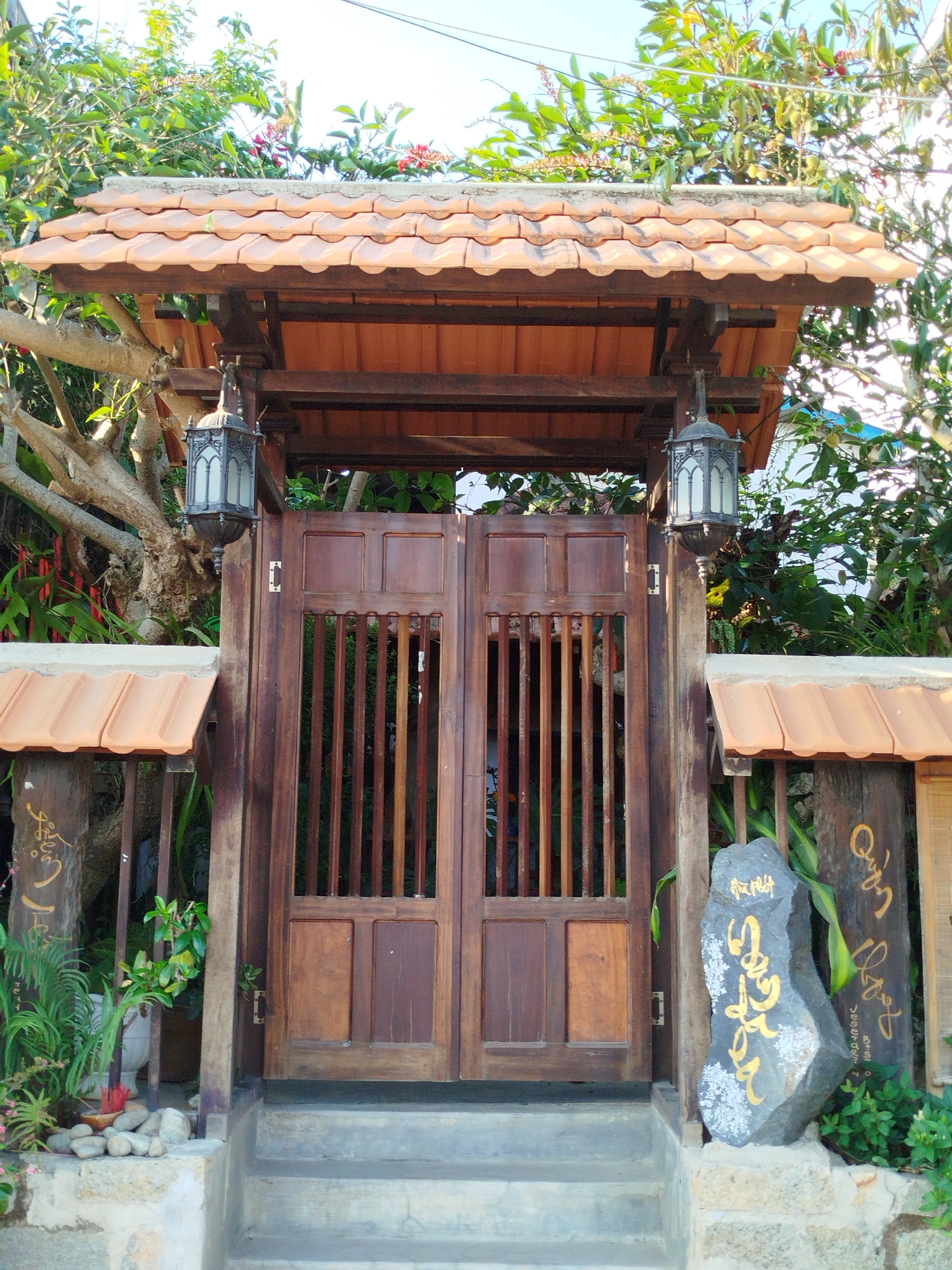 The image depicts a traditional Vietnamese-style wooden gate, possibly the entrance to a residence or business, with architectural details typical of the Da Lat region in Vietnam. The gate is constructed of dark brown wood and has a tiled roof above it. Ornate metal lanterns hang on either side of the gate. The gate itself has vertical slats and solid rectangular panels. To the left of the gate, a wooden post with a tiled roof covers a wooden plaque with Vietnamese script; similar architecture on the right has a stone with Vietnamese script. Plants and greenery surround the gate. Stone steps lead up to the gate, further suggesting a welcoming entrance.