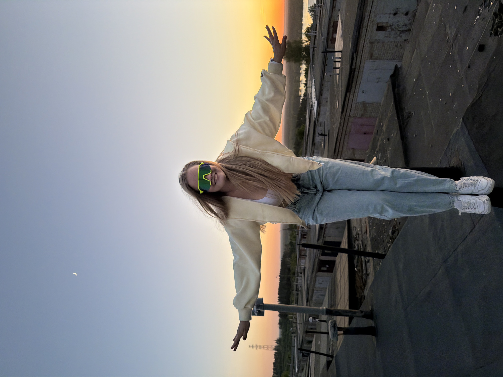 A young woman with long blonde hair wearing light-wash jeans, a white top, a cream-colored zip-up jacket, and bright green sunglasses stands on the edge of a rooftop at sunset. She has her arms outstretched to the sides. The background reveals an urban landscape with industrial buildings and infrastructure typical of an industrial area. The sky is transitioning from a dark blue to orange gradient indicating either sunrise or sunset. A crescent moon is visible in the blue portion of the sky. The location is likely in an urban area similar to Chaikovsky, Russia, given the presence of industrial structures.