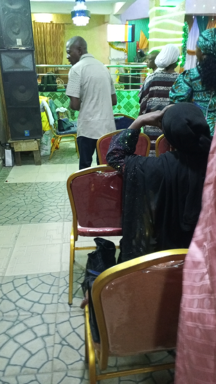 Inside a room with tile flooring, a man in a light shirt walks near a speaker. Several individuals are seated. One wears a white head covering, while another is adorned in green. The person in the foreground dons a black garment and head covering. Rows of chairs with gold frames are visible. Decorations include a chandelier and green streamers. A screen displays a colorful image in the background.