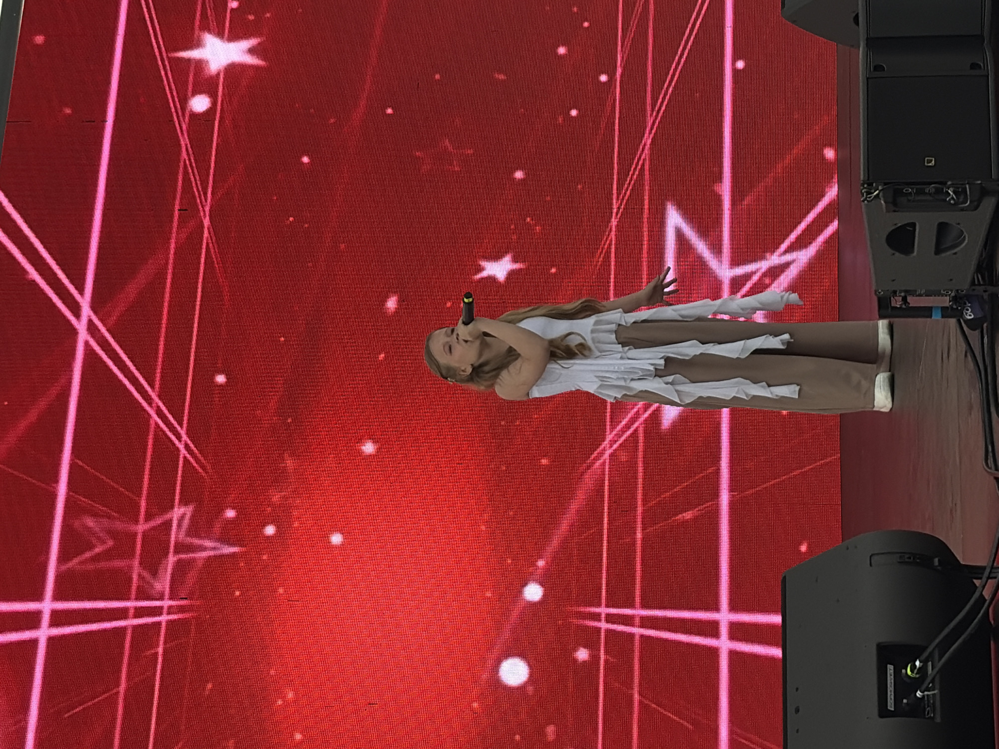 A young girl with blonde hair is singing into a microphone on a stage in front of a large red LED screen. The screen displays a pattern of pink lines and stars, and some white dots. The girl is wearing a white ruffled top and tan pants. She is standing in front of a large black speaker. The image appears to have been taken outdoors during the daytime, as evidenced by the bright lighting. This scene is likely from an event in Izhevsk, Russia, potentially a festival or concert.