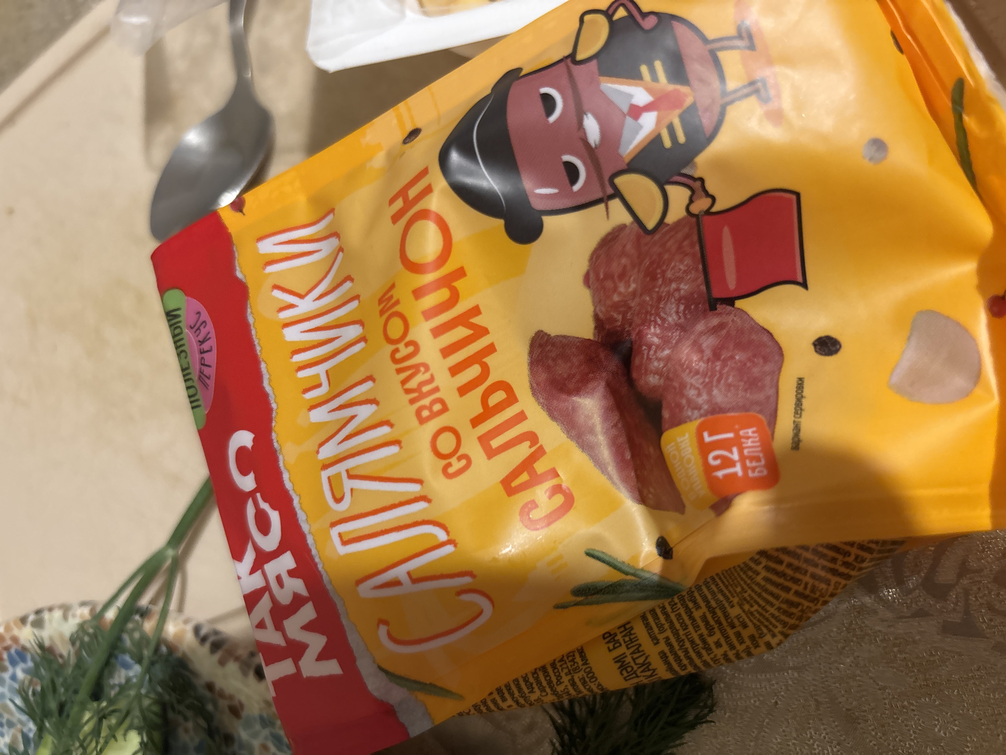 The image shows a close-up of a yellow and red packaged food product. The packaging features large white lettering that appears to be in Russian, with the words "САЛЯМИЧИКИ" (salamichiki) and "СО ВКУСОМ САЛЬЧИЧОН" (so vkusom salchichon) prominently displayed. There is a cartoon illustration of a red, bean-shaped character wearing a black hat and a red suit. Several slices of salami are visible within the packaging. In the background, a silver spoon and a sprig of dill are present. The scene appears to be indoors, with a textured surface in the foreground. There is no information in the image that places it in the city of Sinyavino, Russia.
