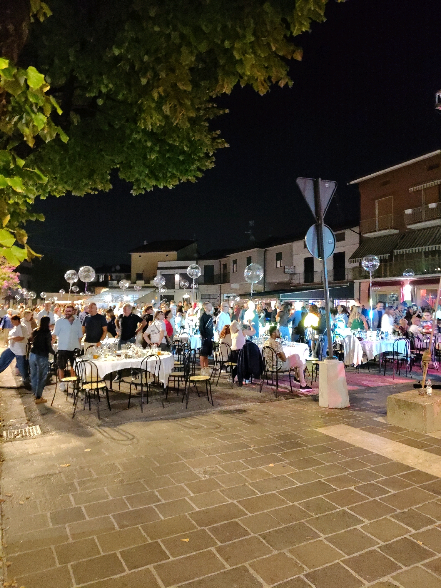 This image depicts an outdoor evening gathering in a city, possibly a street or plaza, as suggested by the cobblestone pavement and buildings in the background. The scene is illuminated by artificial lights, creating a festive atmosphere. Several tables are set up with white tablecloths and chairs, indicating a dining event or celebration. People of various ages are present, some standing and conversing, while others are seated at the tables. There are clear balloons floating above some of the tables, adding to the celebratory ambiance. In the background, the facades of buildings suggest a commercial or residential area. The presence of trees with green foliage indicates a mild climate or season.