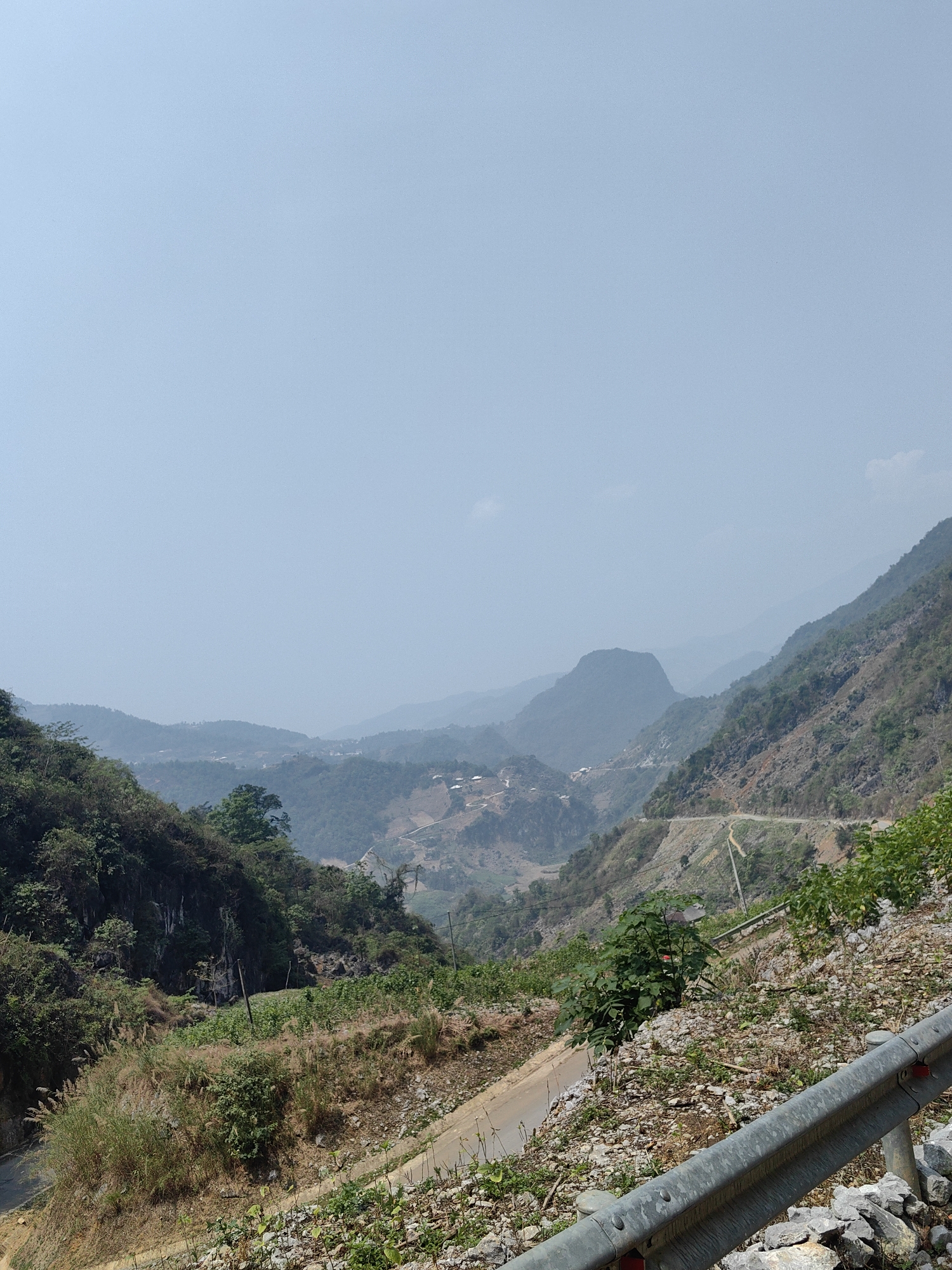 The image depicts a mountainous landscape in Xã Khánh Xuân, Vietnam, under a clear, bright sky. The foreground features a road with a metal guardrail. The hillside adjacent to the road is covered in vegetation, including grasses, shrubs, and small trees, along with rocks and loose soil. In the middle ground, a valley reveals a network of roads and terraced fields. Further in the distance, successive layers of hills and mountains fade into the horizon, creating a sense of depth and vastness. The overall scene is one of rural serenity.