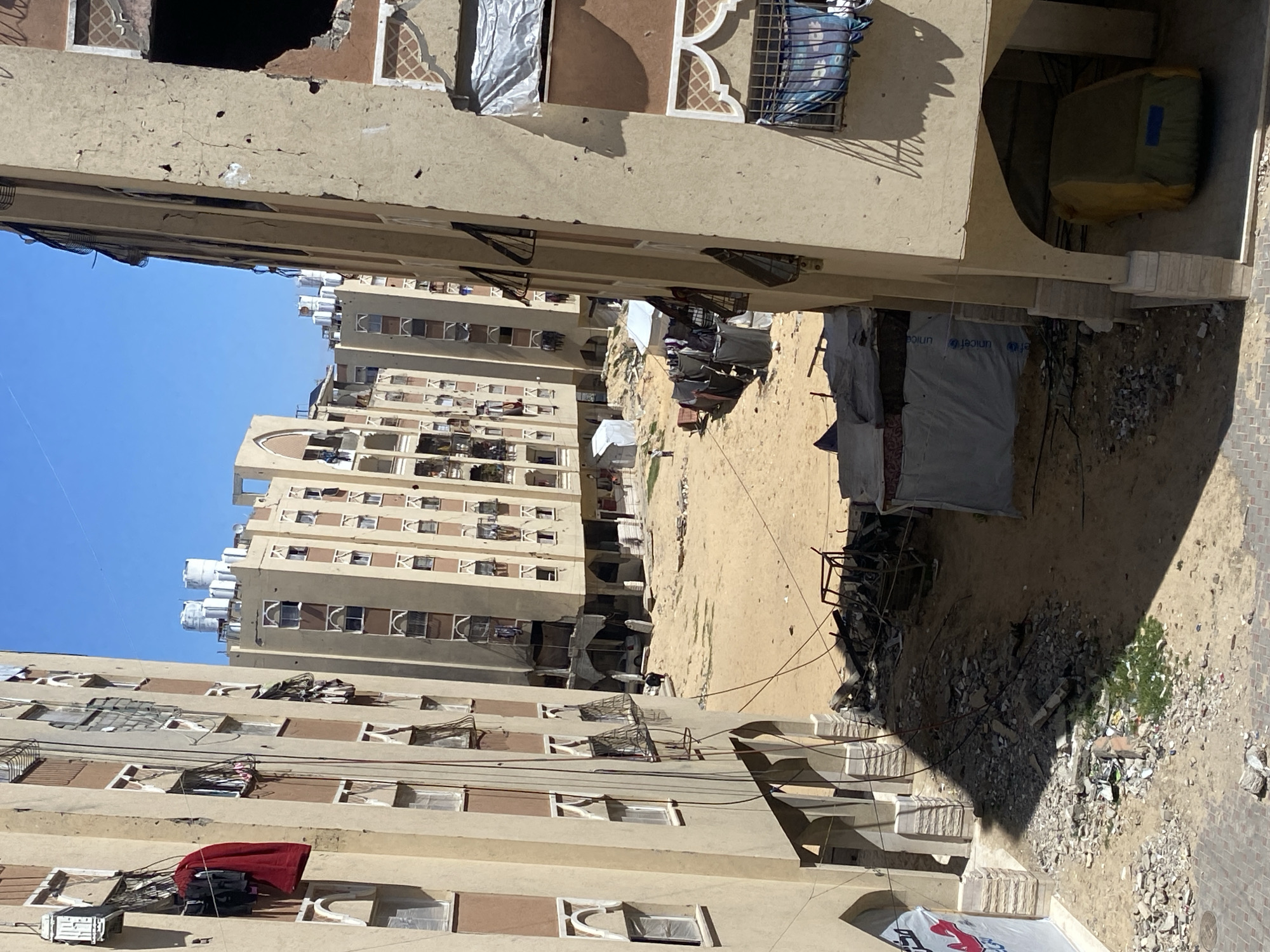 The image depicts a cityscape in Khan Yunis, Palestinian Territory, likely showing damage. The scene is viewed from an elevated perspective, looking down into a courtyard or open area between multi-story buildings. The buildings appear to be residential, with repetitive window designs. Some windows are covered with plastic or cloth. There are objects on the ground, possibly including debris and temporary shelters. There are no visible people in the frame. The sky is visible and clear.