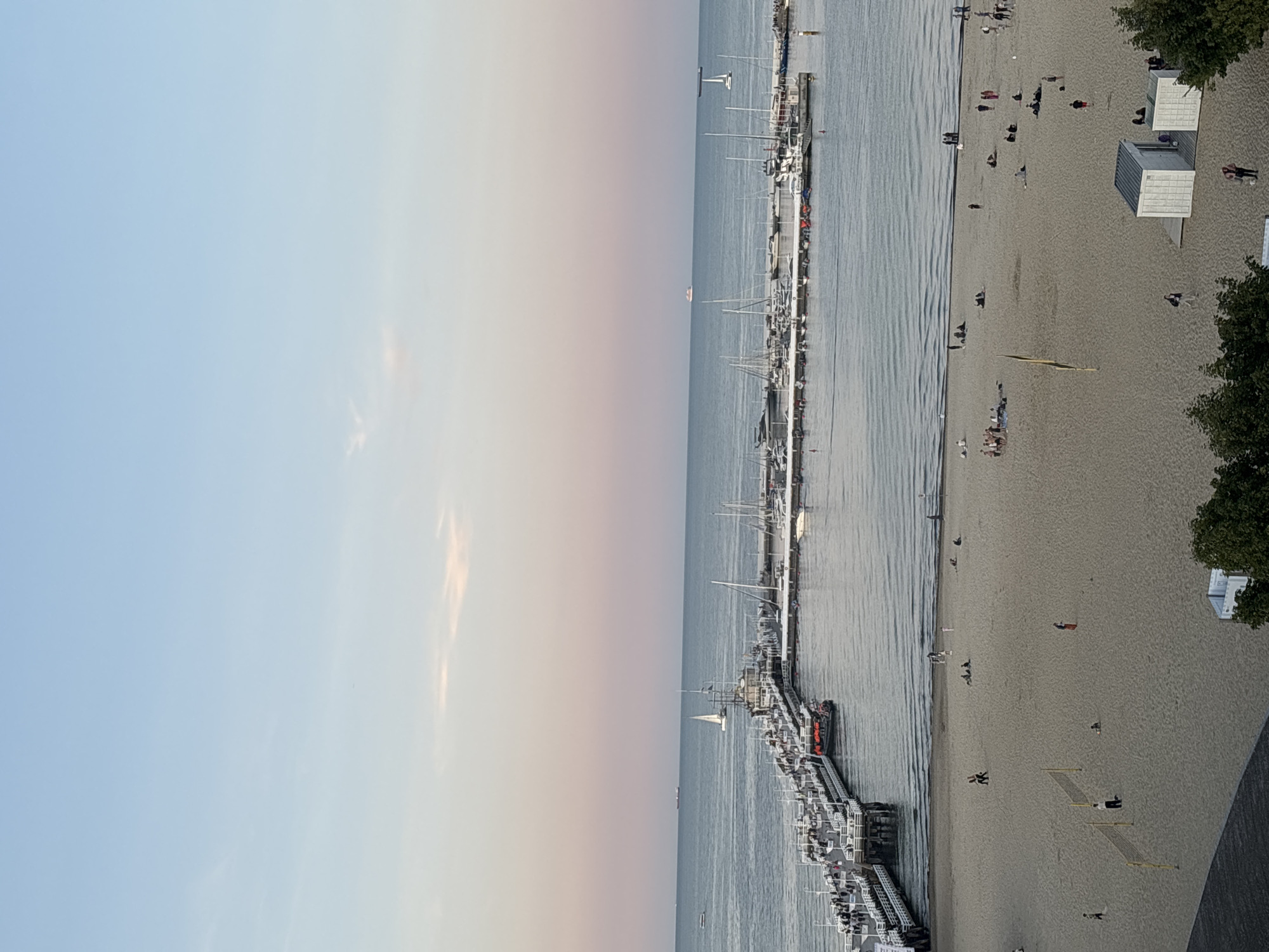 The image shows a view of the Sopot Pier in Sopot, Poland, during what appears to be sunset or sunrise. The sky transitions from a pale blue at the top to soft pink and orange hues near the horizon.
The Sopot Pier, one of the longest wooden piers in Europe, extends into the Baltic Sea. It is lined with people strolling and is adorned with white railings. Several boats, including sailboats and smaller vessels, are docked in the marina adjacent to the pier.
The foreground of the image features a sandy beach with scattered groups of people. Some individuals are sitting or lying on the sand, while others are walking or engaged in activities. Two volleyball nets are set up on the beach, suggesting recreational use. Two small, white changing rooms or beach huts are visible on the right side of the beach.
The sea is calm, with gentle waves lapping at the shore. In the distance, several ships and boats can be seen on the horizon. The overall scene depicts a serene coastal environment with both active and relaxed human presence.