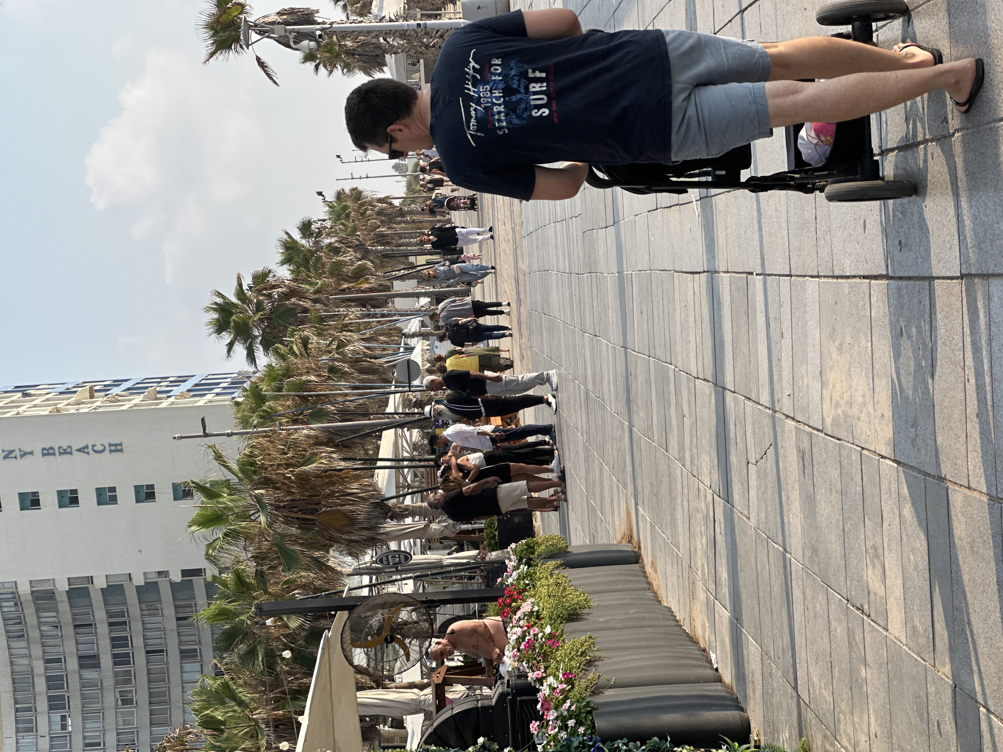 The image depicts a sunny daytime scene on a paved promenade in Bat Yam, Israel, with the Mediterranean Sea likely to the left and a modern high-rise building to the left background. A man wearing a dark blue t-shirt with "Tommy Hilfiger" and "SURF" printed on it and light grey shorts pushes a stroller from behind. He is walking on the promenade, away from the viewer, towards a line of palm trees. Several other people are visible in the background, walking and conversing. To the left, there's an outdoor seating area with umbrellas and a partial view of a building facade with the words "NY BEACH" visible vertically. The promenade is paved with large, rectangular stones, and shadows from palm trees and people are cast on the ground.