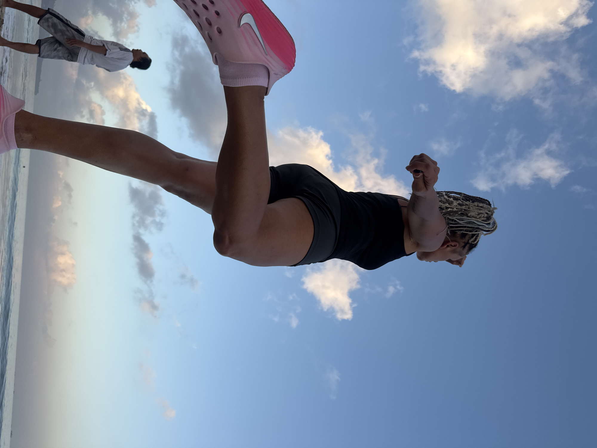 The image depicts a low-angle shot of a woman with blonde dreadlocks in mid-stride on a beach, likely at sunrise or sunset given the sky. She is wearing a black tank top and grey athletic shorts, with pink and silver running shoes. Her left leg is extended forward in a running motion. In the background, a man in a white t-shirt and patterned shorts stands near the water's edge, looking out at the ocean. The ocean is calm with gentle waves, and the sky is a gradient of blue with scattered white and grey clouds. The location is identified as Kuta Selatan, Indonesia.