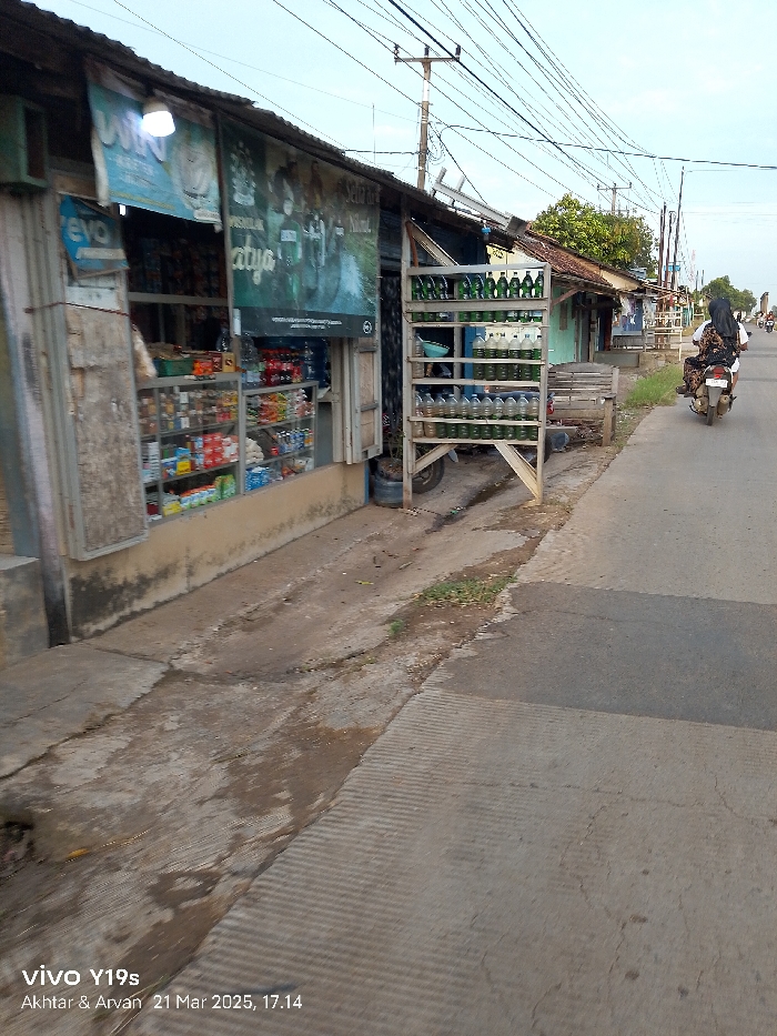 The scene is a narrow, paved alleyway in Anjatan, Indonesia. The alleyway is lined with small, simple buildings, one of which is a shop with a glass front. The shop has various products displayed on shelves inside, including bottles of drinks, packaged food, and other miscellaneous items. There is an advertisement for a motorcycle brand on the side of the shop, next to a sign with the words "Saya" and "Atyo" printed on it. There are also many power lines running across the alleyway. A metal rack full of green and clear plastic bottles stands in front of the shop. There is a person riding a motorcycle in the distance, and some plants are growing in the cracks of the pavement. The alleyway is empty except for the rider and the motorcycle. The time is approximately 5:14 PM.