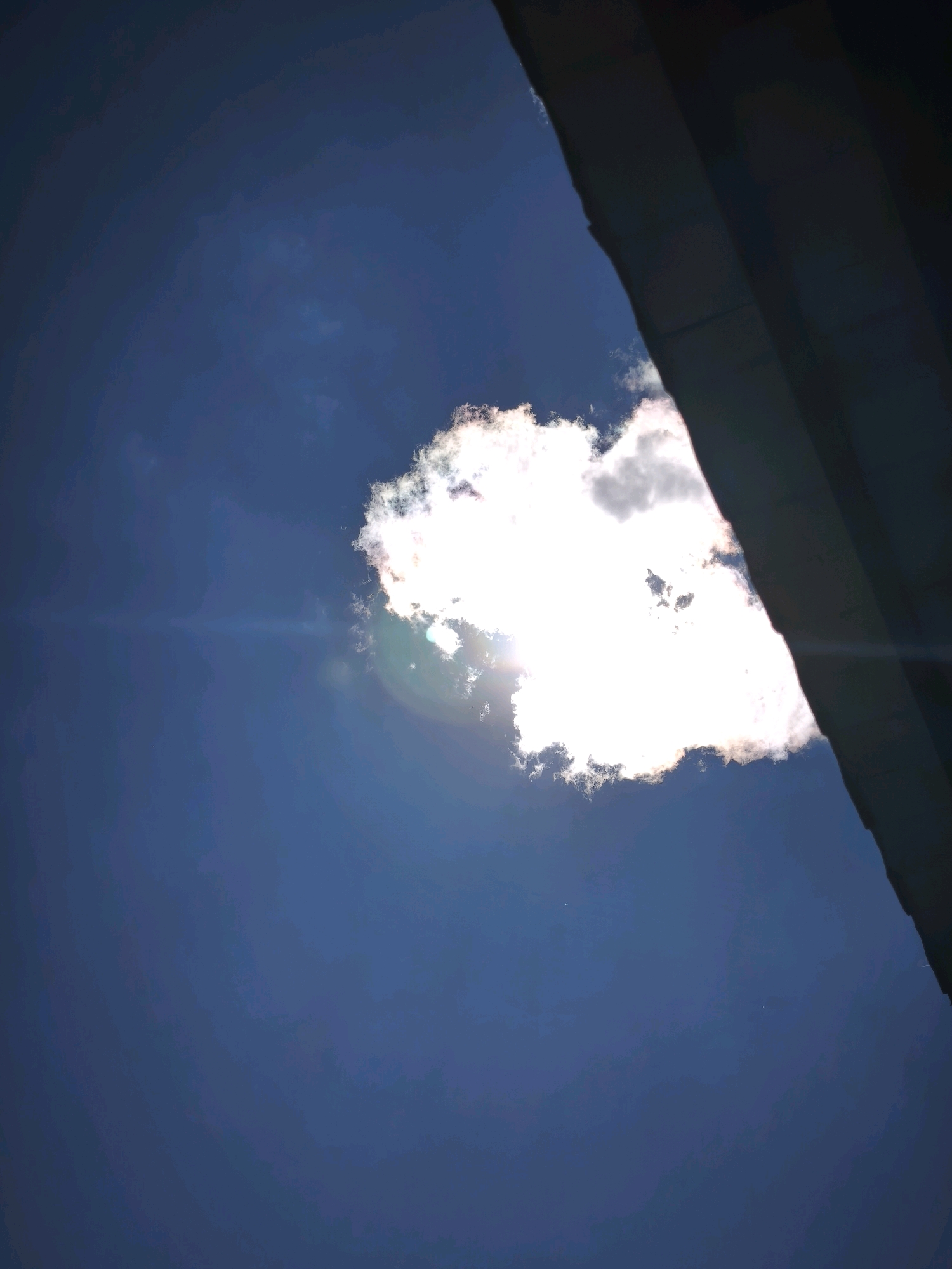 The image shows a dark, angular structure in the upper right corner, obscuring a portion of the sky. A bright, fluffy cloud with the sun shining behind it dominates the center of the frame, casting a lens flare across the deep blue sky. The cloud is illuminated from within, with its edges appearing wispy and diffused. The overall impression is that of a bright day with a single cloud partially covering the sun, viewed from a low angle, possibly beneath an overhang. The location in Karachayevsk, Russia is not discernible from the image itself.