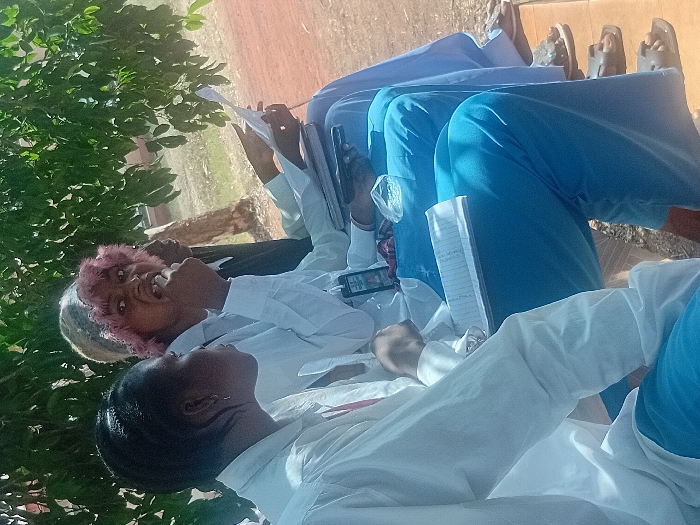 Several people sit outdoors beneath the shade of a tree. They are dressed in white shirts and blue pants or skirts. Some have notebooks and papers in their hands, while others hold cell phones. One person has pink hair and another wears a patterned cap. The group seems to be focused on reading or writing, possibly in a class or meeting. Some sandals are visible at the edge of the frame.