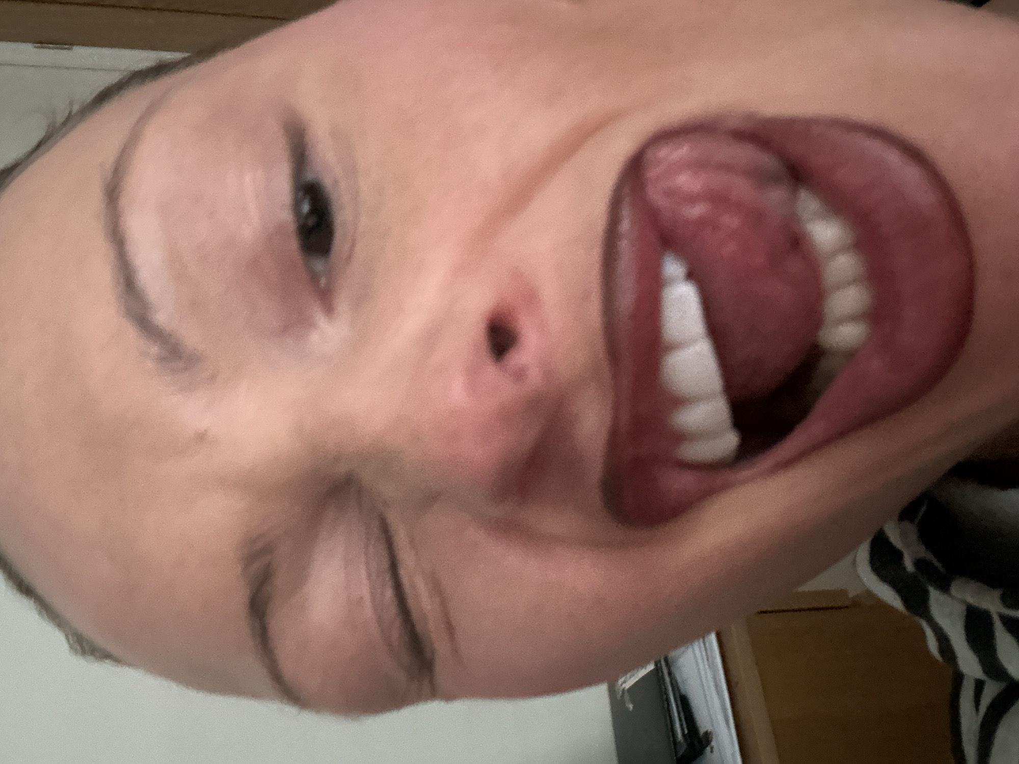 A close-up, front-facing image depicts an individual, likely female, in an indoor setting located in Wealden, United Kingdom. The subject's face fills the frame, presenting a playful expression: her left eye is closed in a wink, while her right eye remains open. Her mouth is wide open, exposing white teeth and a protruded tongue. The lips are colored with a dark red or burgundy lipstick. The skin appears light-toned, with minor facial creases around the winking eye. The background is largely out of focus, revealing a light-colored wall on the upper left, an indistinct dark object resting on papers atop a light brown wooden surface in the lower left, and a partially visible black and white striped garment at the bottom of the frame.