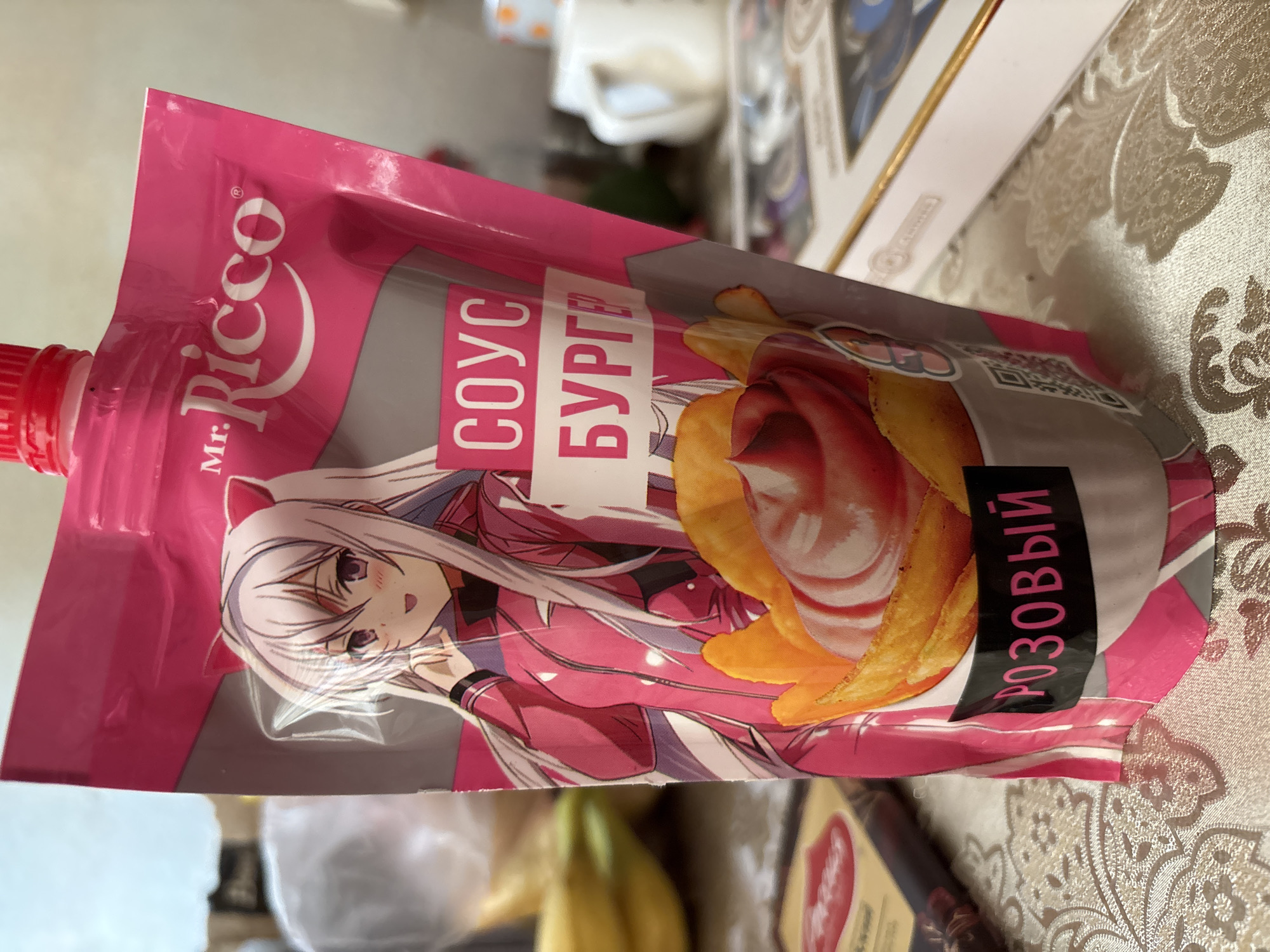 The image shows a pink pouch of "Mr. Ricco" burger sauce. The pouch features an anime character with long white hair and pink cat ears, wearing a pink and black outfit. The text on the pouch is in Russian and says "СОУС БУРГЕР" (Sauce Burger) and "РОЗОВЫЙ" (Pink). There is also a picture of the sauce drizzled over potato chips. The pouch is sitting on a table covered with a patterned tablecloth. In the background, out of focus, there are bananas, a white mug, and a box. The lighting suggests an indoor setting, possibly a kitchen or dining area. The image does not contain any information about the specific location in Saint Petersburg, Russia.