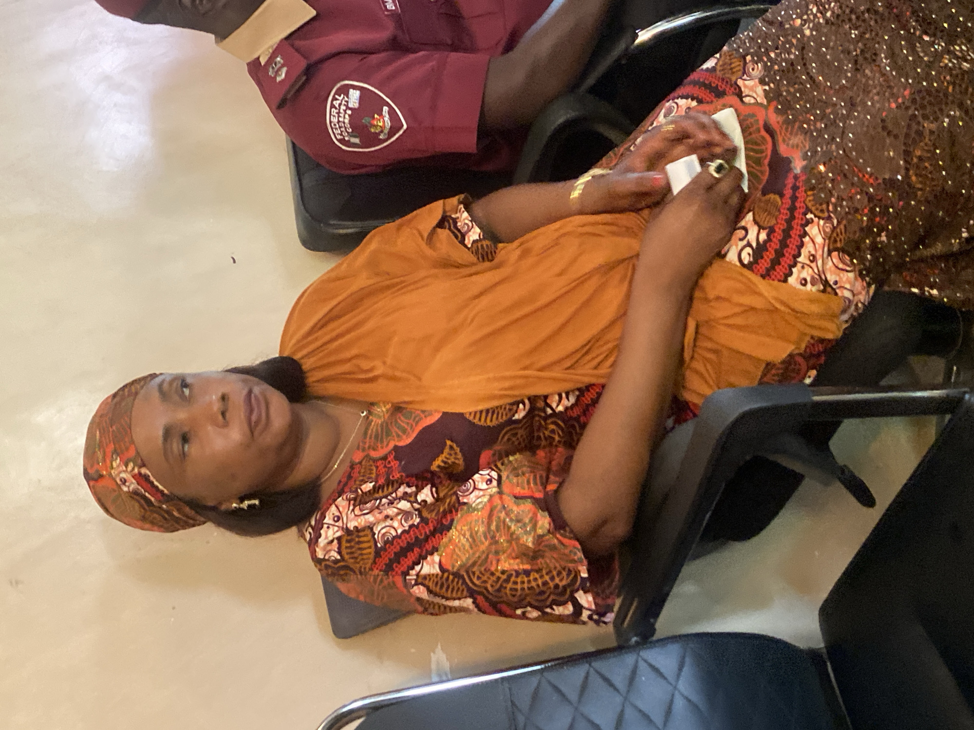 In Unguwar Sarki, Nigeria, an image captures a seated woman from the chest up, her attention directed to her right. She is adorned in a vibrant, patterned outfit featuring rich orange, brown, and red hues, accentuated by sequined embellishments on the lower portion of her garment. A matching headscarf frames her face. To her right, a man in a maroon uniform with tan accents is partially visible, his arm resting on the chair. The setting appears to be indoors, with a neutral-colored wall behind them. The woman holds a small white object in her hands, one of which displays a ring with a dark stone.