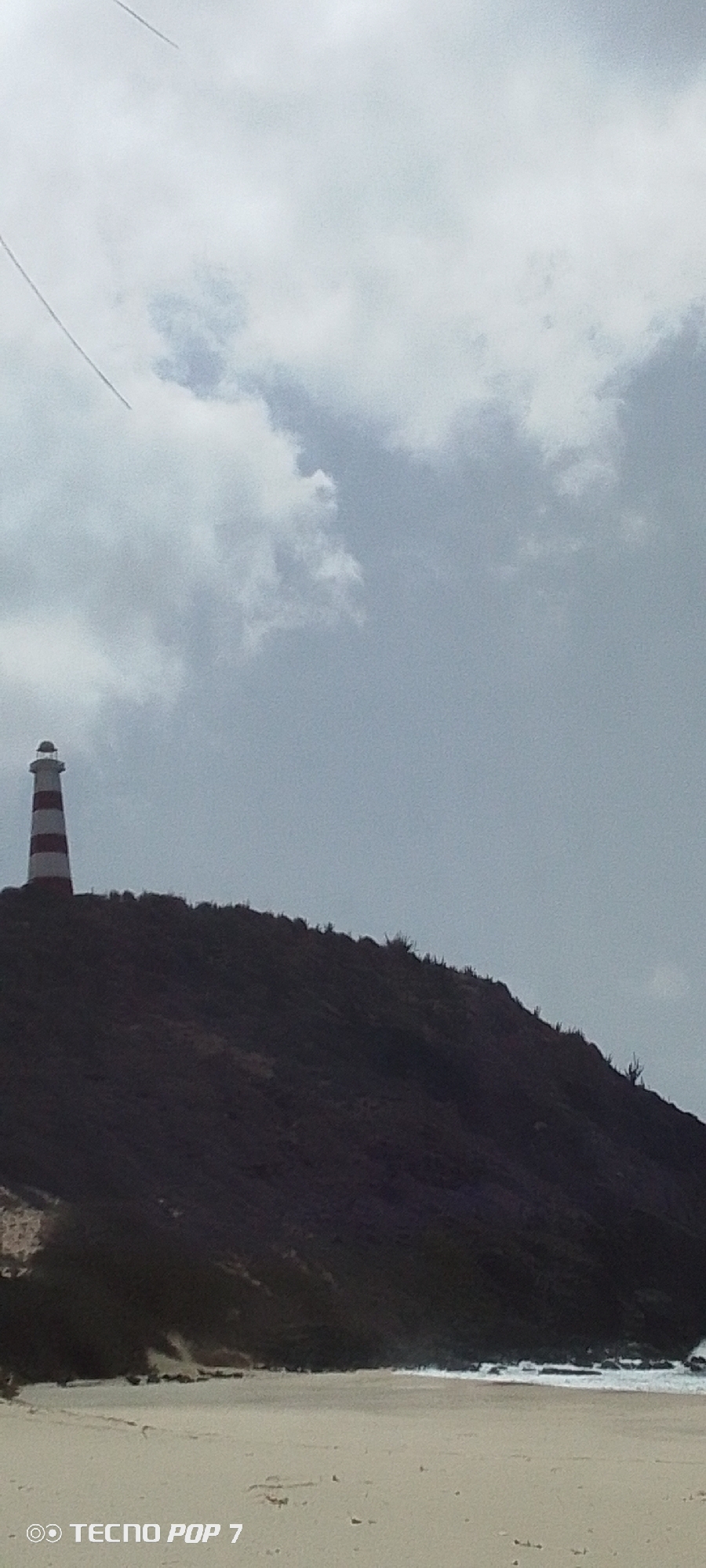 The image depicts a coastal scene on Margarita Island, Venezuela, specifically in Pedro González. A red and white striped lighthouse stands prominently atop a dark, vegetation-covered hill. Below the hill, a sandy beach stretches to the water's edge where small waves break. The sky is overcast with gray clouds. There is a smartphone watermark located in the bottom left corner.