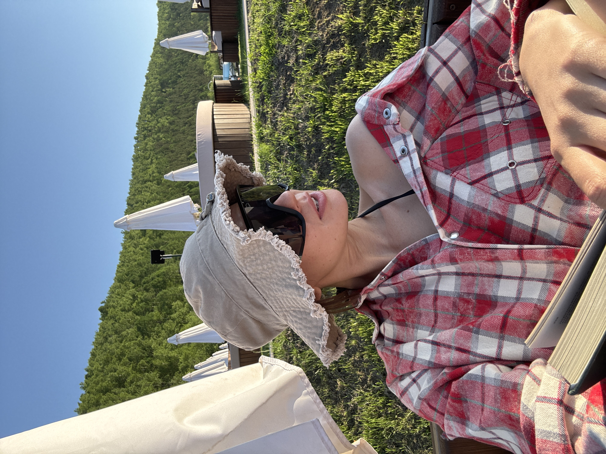 The image depicts a person relaxing outdoors on a bright, sunny day, captured from a slightly elevated perspective. The subject, likely a woman, is partially visible, wearing a red, white, and grey plaid shirt unbuttoned over a black bikini top. She has dark sunglasses perched on her face and a light beige bucket hat with a frayed brim. A book with a dark cover and light pages is held in her lap or hands, suggesting she is reading.
The background reveals an outdoor leisure area, possibly a resort or recreational spot. Several white sun umbrellas are visible, along with wooden outdoor lounge chairs arranged on a vibrant green lawn. Beyond this, a densely forested hillside or mountain range rises under a clear blue sky. The strong, direct sunlight indicates it is daytime, likely mid-day or afternoon, and the weather is fair and warm. The person appears relaxed and content, with a slight smile. The overall atmosphere is one of leisure and tranquility in an outdoor setting, consistent with a recreational area in Дрокино, Russia. No visible text is discernible in the image.