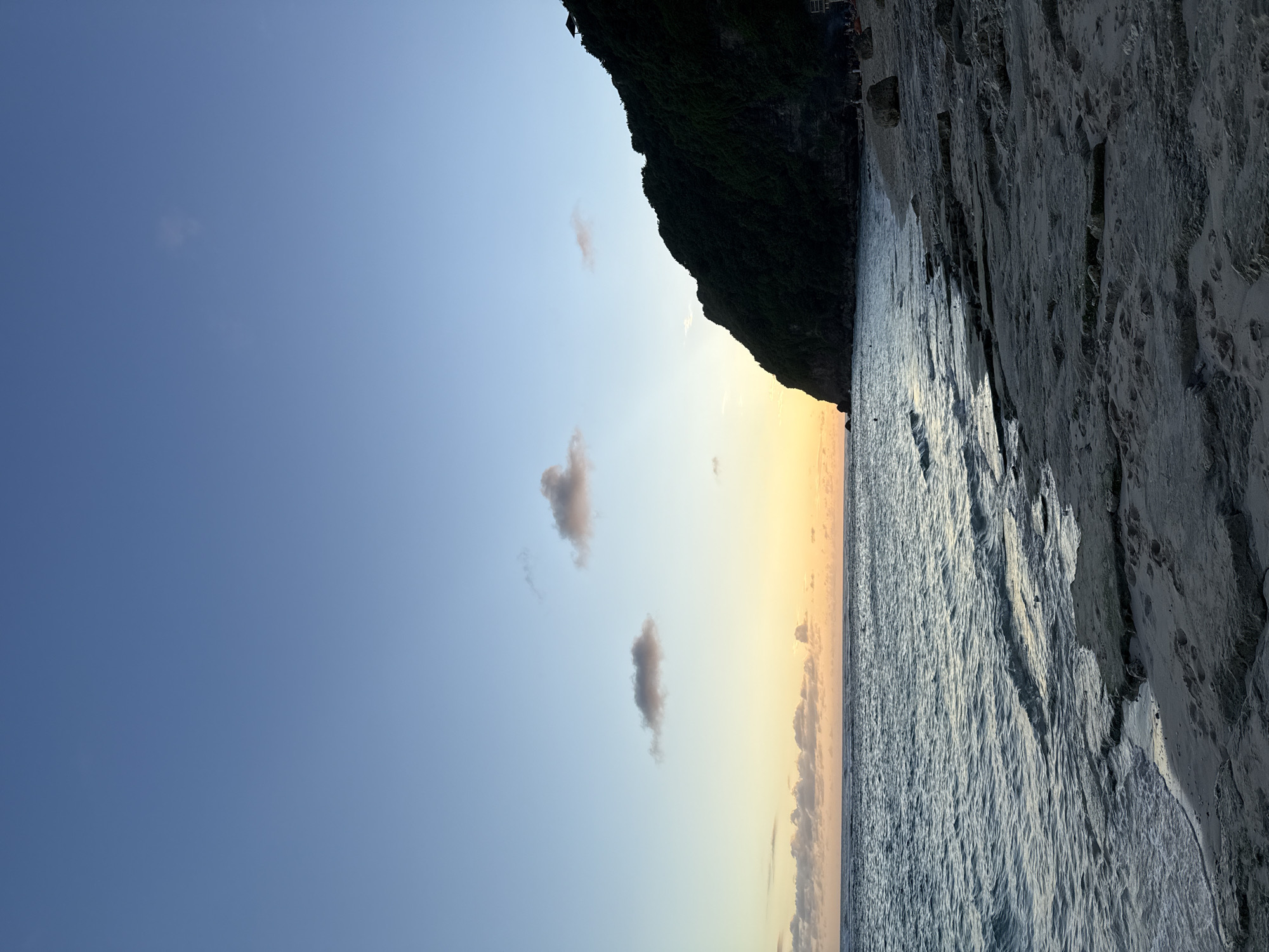 The image shows a beach scene in Ungasan, Indonesia. The view is split between the sky and the sea. The sky, transitioning from blue to a yellow sunset near the horizon, contains sparse clouds. The sea reflects the sunset's colors. A cliff face covered in dark green vegetation dominates a part of the horizon. To the right is a rocky or sandy section of the beach. The image is oriented vertically.