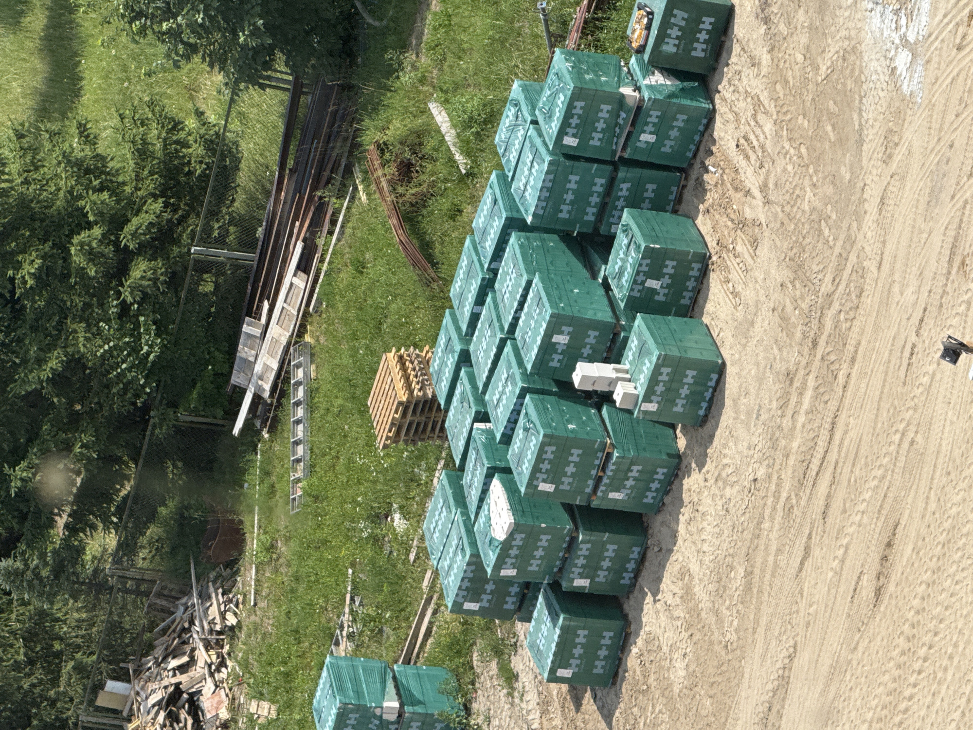 The image, taken from an aerial perspective in Mikołajki, Poland, shows a construction site or storage area on a sunny day. The primary focus is a large stack of green-wrapped building materials, likely blocks or bricks, organized neatly on the ground. Each package displays the "H+H" logo prominently. The surrounding area includes lush green grass and trees, suggesting an outdoor, possibly rural or semi-rural, setting. In the background, there are piles of lumber and metal, along with fencing and a ladder, indicating ongoing construction or demolition work. The ground is a mix of dirt and sand with visible tire tracks, reinforcing the idea of a work-in-progress environment. There are no people visible in the image, and the overall impression is one of a functional, outdoor workspace.