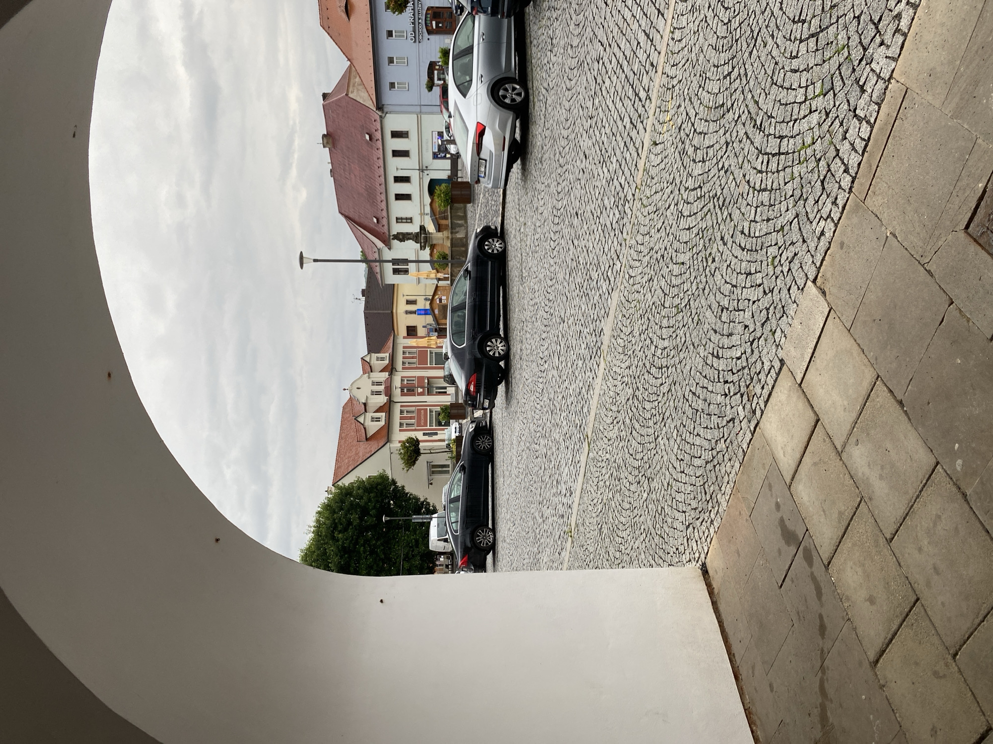 The image depicts a view through a large white archway onto a cobblestone plaza in Bílina, Czechia. Several cars are parked on the plaza, including a silver sedan and two dark sedans. Buildings with red tiled roofs line the plaza, some featuring ornate facades and windows. A street lamp is visible in the distance. The sky is overcast with clouds. The foreground shows large square paving stones leading up to the archway.