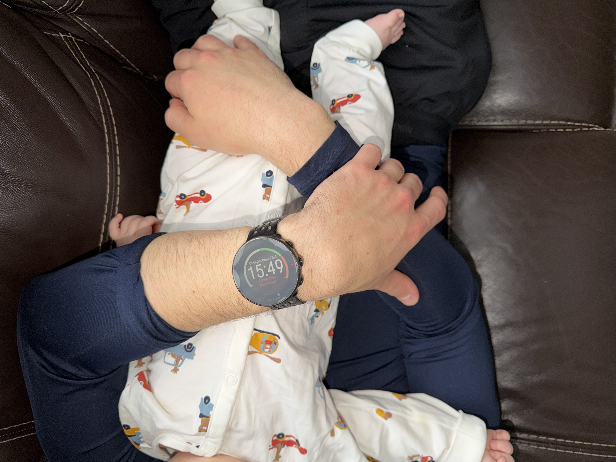 A person is holding a baby in their lap while seated on a brown leather couch. The person is wearing a long-sleeved navy blue shirt and dark blue pants. The baby is wearing white pajamas with a cartoon vehicle pattern. The person is wearing a black smartwatch on their left wrist that displays the time as 15:49 and the date as Sunday, April 26th. The watch face is set to the Russian language. The environment appears to be indoors, possibly a living room. There is no information to indicate the location as Khimki, Russia, or any city in Russia.