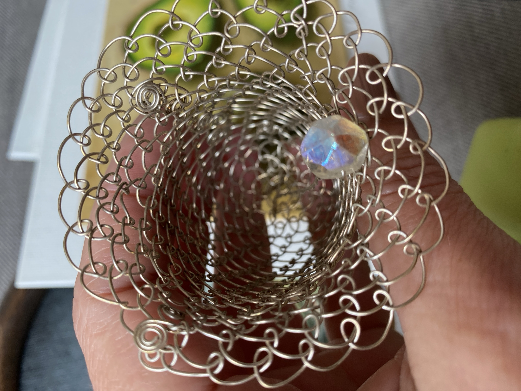 A close-up, top-down shot shows a person's hand holding a decorative, silver wire-mesh cylindrical object. The object is open at both ends and has an intricate, looped pattern. Inside the cylinder, near the bottom, a faceted, iridescent bead is visible. The background is blurred but suggests an indoor setting with what appear to be slices of avocado in the upper right quadrant. No specific location in Bílina, Czechia can be determined from the visual information provided.