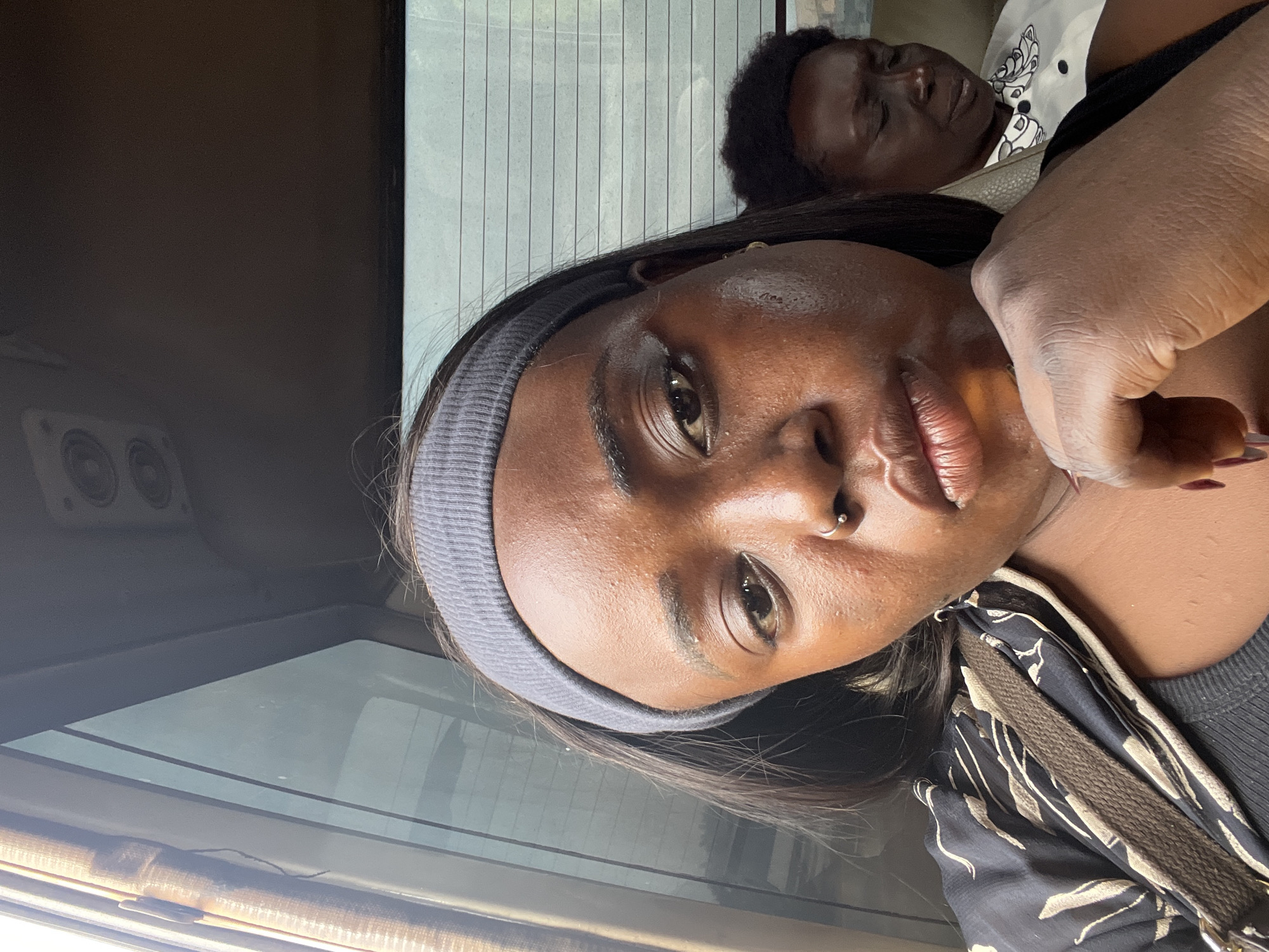 In Kugbo, Nigeria, a dark-skinned woman is photographed from the chest up, leaning on her hand. She is wearing a grey headband and a black patterned top. Her eyes are partially closed and she has a nose ring. Behind her, another woman is visible with her eyes closed, wearing a white shirt with a black collar and a patterned detail. The background appears to be the interior of a vehicle, with a window on the left and a dark ceiling with speakers visible at the top.