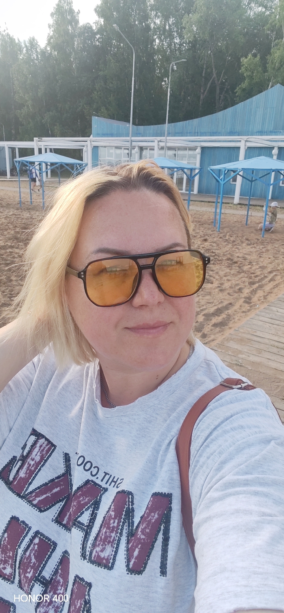 A woman with blonde hair and sunglasses is taking a selfie on a sandy beach. She is wearing a light-colored t-shirt with a graphic print and a brown strap from a bag is visible across her shoulder. The background shows a beach with several blue canopies and a wooden walkway. In the distance, there are trees and a blue building. The scene appears to be a recreational area. The lighting suggests it is daytime, likely late afternoon due to the warm tones. The image has a casual and relaxed feel. Visible text on the t-shirt appears to be part of a graphic design. The bottom left corner shows "HONOR 400".