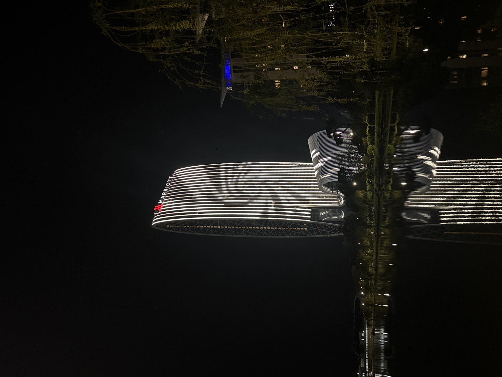 The image shows the Batumi, Georgia Marriott Hotel at night, reflected in the water, inverted. The hotel is illuminated with horizontal white lines, with a red "Marriott" logo visible. There are some trees visible between the Hotel and its reflection in the water. In the top right, another building is visible.