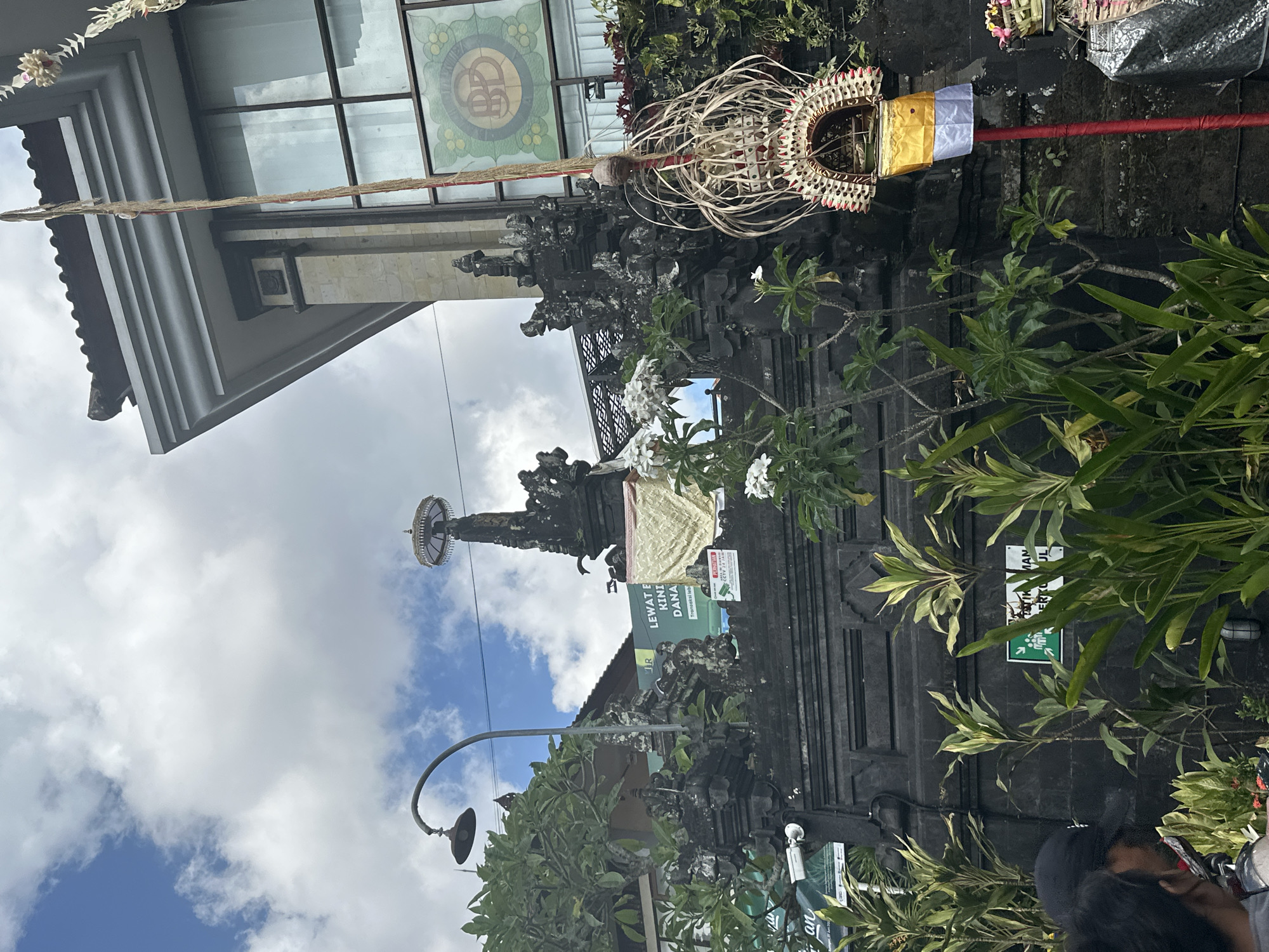 In Ubud District, Indonesia, this image captures a street-level view of a stone wall with ornate carvings, possibly part of a temple or traditional Balinese architecture. A prominent feature is a tall, red pole adorned with intricate woven decorations and a golden and white flag, suggesting a ceremonial or cultural event. Lush green foliage and plants surround the wall, with white flowers blooming in the foreground. In the background, a modern building with large windows and a distinct circular logo is visible, contrasting with the traditional elements. The sky above is partially cloudy, with patches of blue. A street lamp extends from the left side of the frame, and a security camera is mounted on the wall. Two individuals are partially visible in the lower left corner, their faces obscured. Several signs are posted on the wall, one in green with figures and text, another with Indonesian text. The overall scene suggests a blend of traditional and modern life in a vibrant cultural setting.