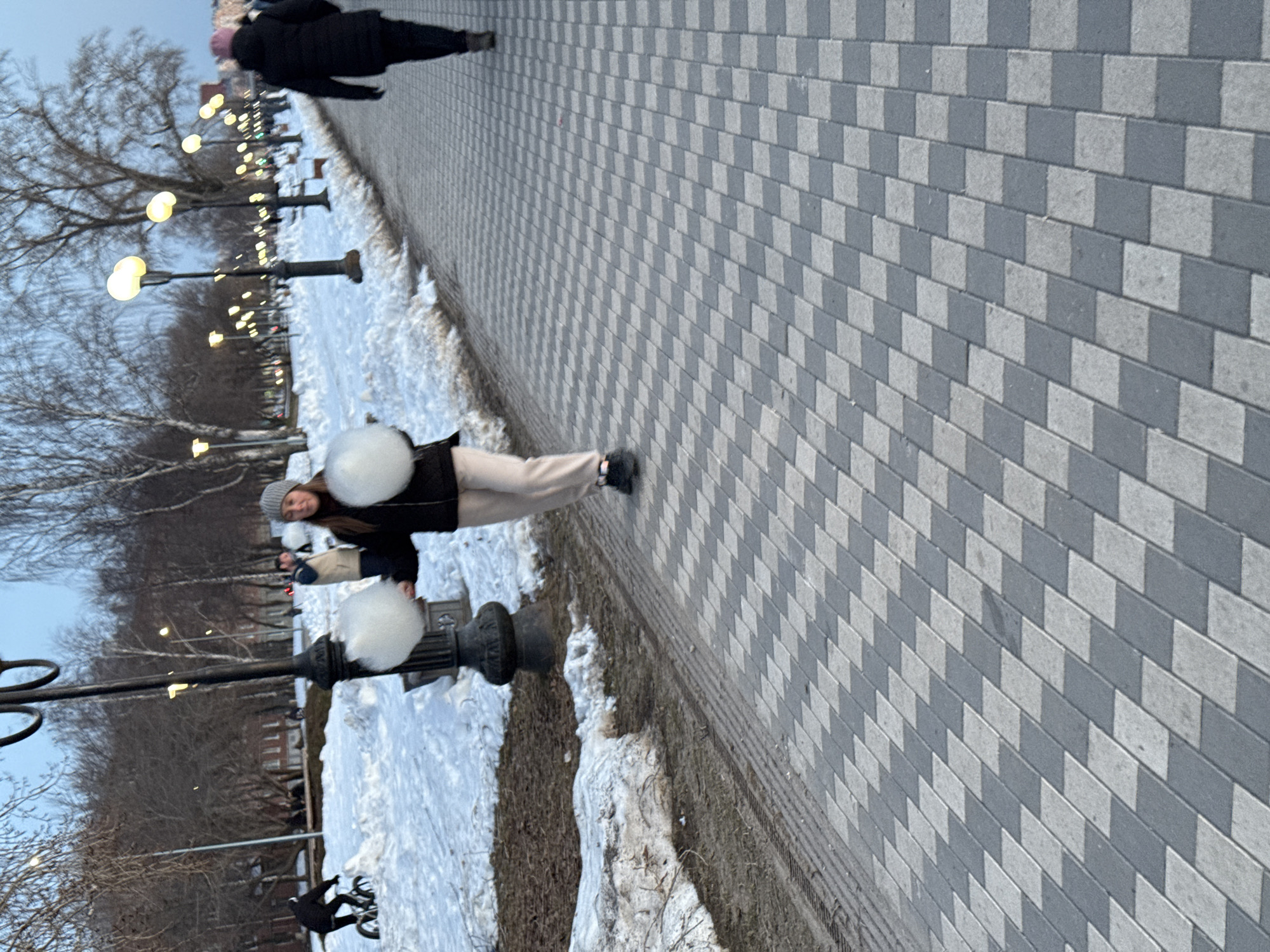 The image depicts a street scene in Votkinsk, Russia, in what appears to be winter. The ground is partially covered with snow. A woman wearing a hat, dark jacket, and light-colored pants is standing on a sidewalk made of alternating light and dark gray paving stones. She is facing the camera. A row of street lamps with spherical lights lines the path, extending into the distance. Bare trees stand along the side of the walkway. In the distance, another person dressed in dark clothing is walking away from the camera. A cyclist is visible in the foreground.