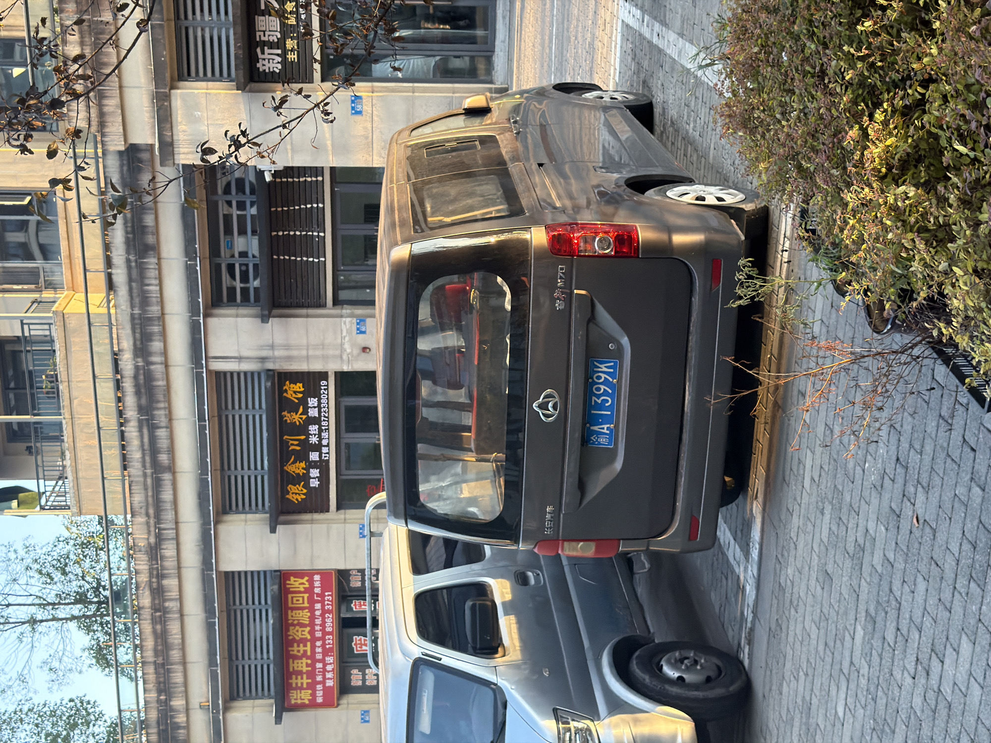 A dark gray minivan is parked in a lot. The minivan has a license plate that reads "渝A 1399K". A silver van is parked to the left of the minivan. In the background, there are buildings with signs in Chinese. One sign reads "瑞丰再生资源回收", and another reads "银鑫川菜馆". The ground is paved with gray bricks. There is a tree with bare branches and autumn leaves in the foreground. The scene appears to be in Jiangjin, China, given the "渝A" on the license plate, which is the vehicle registration code for Chongqing municipality, of which Jiangjin is a district.