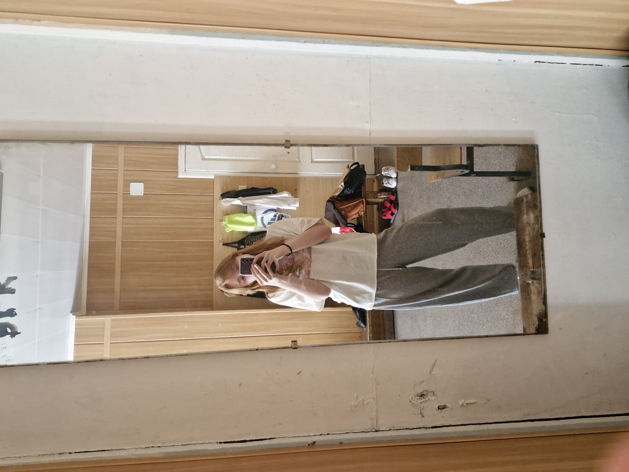 A person stands in front of a large rectangular mirror, taking a selfie with a black smartphone. The person is wearing a light-colored t-shirt and gray sweatpants. The mirror reflects a room with light-colored walls and wooden panels. In the background, there is a white door, storage shelves, and various items including bags, shoes, and clothing. The location cannot be determined from the image.