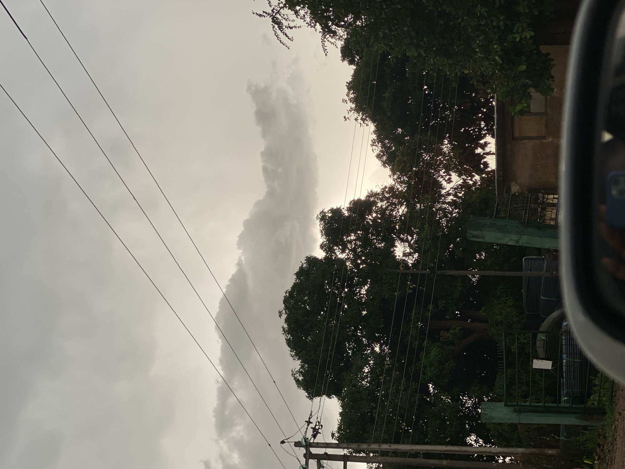 The image shows an outdoor scene in Maigizo, Nigeria, under an overcast sky with thick clouds. Power lines stretch across the sky, running from the lower left to the upper right. A utility pole is visible at the bottom. Trees with dense foliage are situated on the right, partially obscuring a building with a window and a fence. Part of a vehicle is visible on the right edge of the frame.