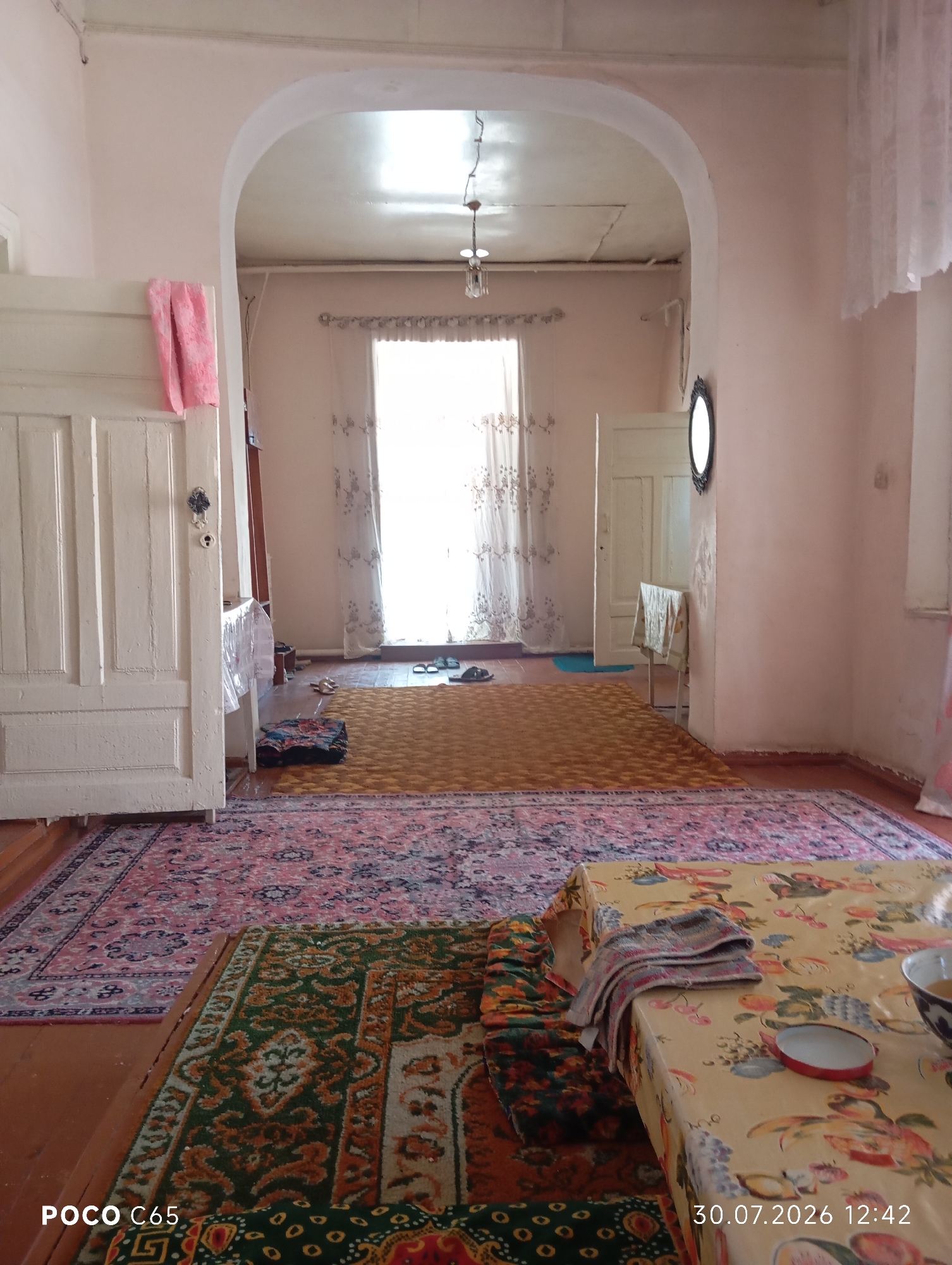 The image depicts the interior of a room in Beruniy, Uzbekistan. The room appears to be a living area or hallway, with multiple rugs covering the wooden floor. A large archway frames the central part of the room, leading to a doorway on the right.
In the foreground, a patterned rug with green and orange motifs is visible, partially obscured by a table covered with a floral-patterned tablecloth. A stack of folded fabric and a small red-trimmed dish sit on the tablecloth.
Beyond this, a larger pink and blue patterned rug stretches across the floor. A yellow rug is laid over part of the pink rug.
To the left, a white wooden door with decorative paneling is prominent. A pink cloth is draped over its top edge.
In the background, a large window with sheer curtains is the primary light source, illuminating the room. Several pairs of sandals are placed near the window. A white door is visible through the archway on the right, and a small table with a patterned cloth stands near it. A dark, oval-shaped mirror hangs on the wall to the right of the doorway. The walls are painted a light pink color. The ceiling is visible through the archway and appears to be textured.