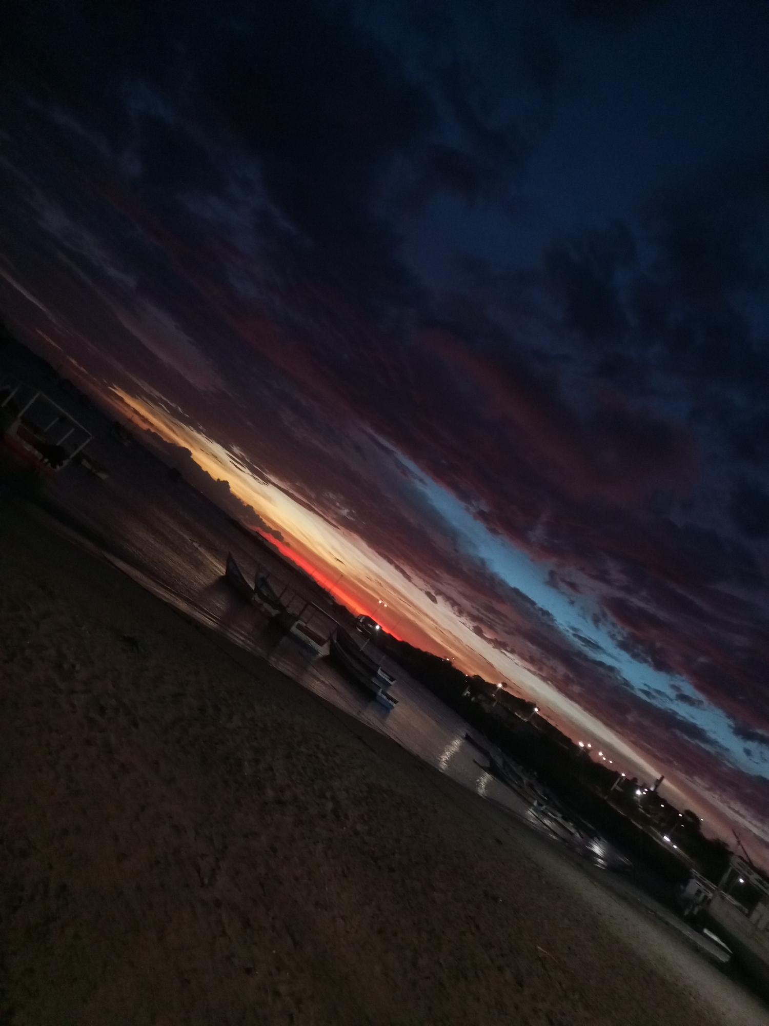 The image depicts a twilight scene at a beach in Juan Griego, Venezuela. The sky is filled with dark, layered clouds, transitioning from dark blue to shades of purple and red near the horizon. A vivid sunset is visible, casting an orange and red glow across the sky and reflecting on the water's surface. Several small boats are moored in the shallow water along the shoreline. The beach consists of dark sand in the foreground, and a line of lights can be seen in the distance, suggesting a town or developed area. The photograph is taken at an angle, which enhances the dramatic effect of the sky and the receding shoreline.