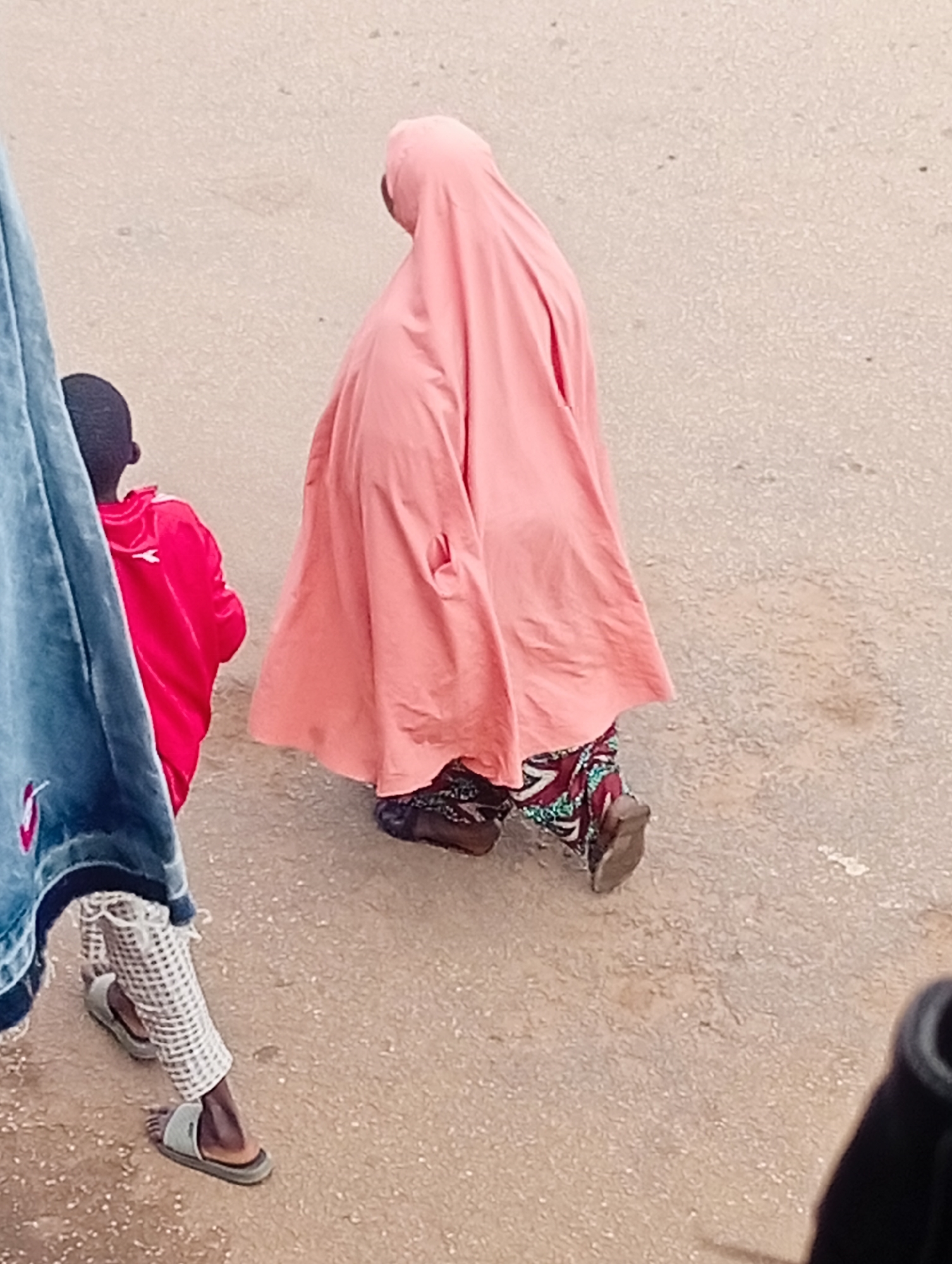 In Bukuru, Nigeria, a woman wearing a salmon pink hijab and a patterned skirt walks on a dirt road, her feet clad in dark, closed-toe shoes. To her left, a young boy in a bright red jacket walks ahead. The scene is framed by a portion of blue denim clothing on the left side of the image. The ground is composed of light brown dirt, with some scattered pebbles.