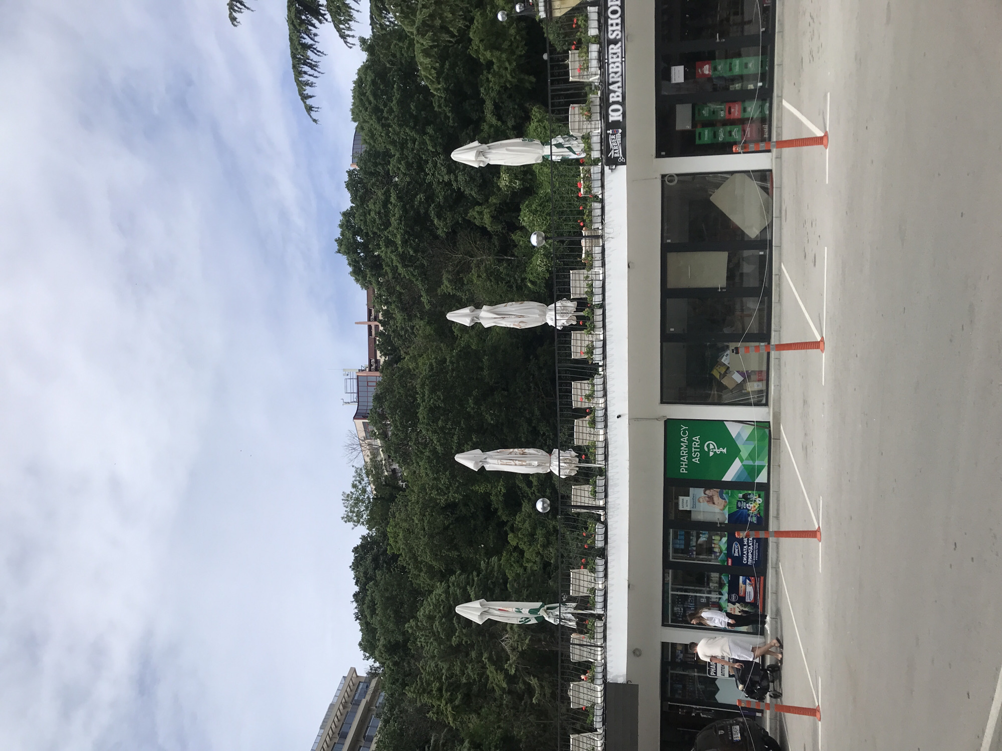 The image shows a commercial strip with a row of businesses and a paved parking lot in front. In the foreground, there are orange bollards and white parking lines on the asphalt. Behind the bollards, the commercial building has large glass windows and doors, indicating shops. One shop is identified as "PHARMACY ASTRA" with green signage. Another shop has signage that reads "IO BARBER SHOP". Several stacks of green and red crates are visible inside the barber shop.
Above the shops, there is a balcony or terrace area with several white umbrellas, some closed and some partially open. A metal railing runs along the edge of this elevated area. Potted plants with red flowers are placed along the railing. A spherical lamp is also present on this level.
Behind the commercial building and the terrace, a densely wooded hillside rises steeply. The trees are lush and green, suggesting a temperate climate. In the background, the upper floors of buildings with balconies and windows are visible peeking through the trees, indicating an urban or semi-urban setting. The sky is overcast with light gray clouds.
In the parking lot in front of the pharmacy, a man in a white t-shirt and shorts is pushing a stroller. A woman, also in a white top, stands beside him. A dark-colored car is parked to the left of the frame. The overall impression is a commercial area nestled against a natural hillside, likely in Varna, Bulgaria, given the presence of a "Pharmacy Astra," a common pharmacy chain in Bulgaria.