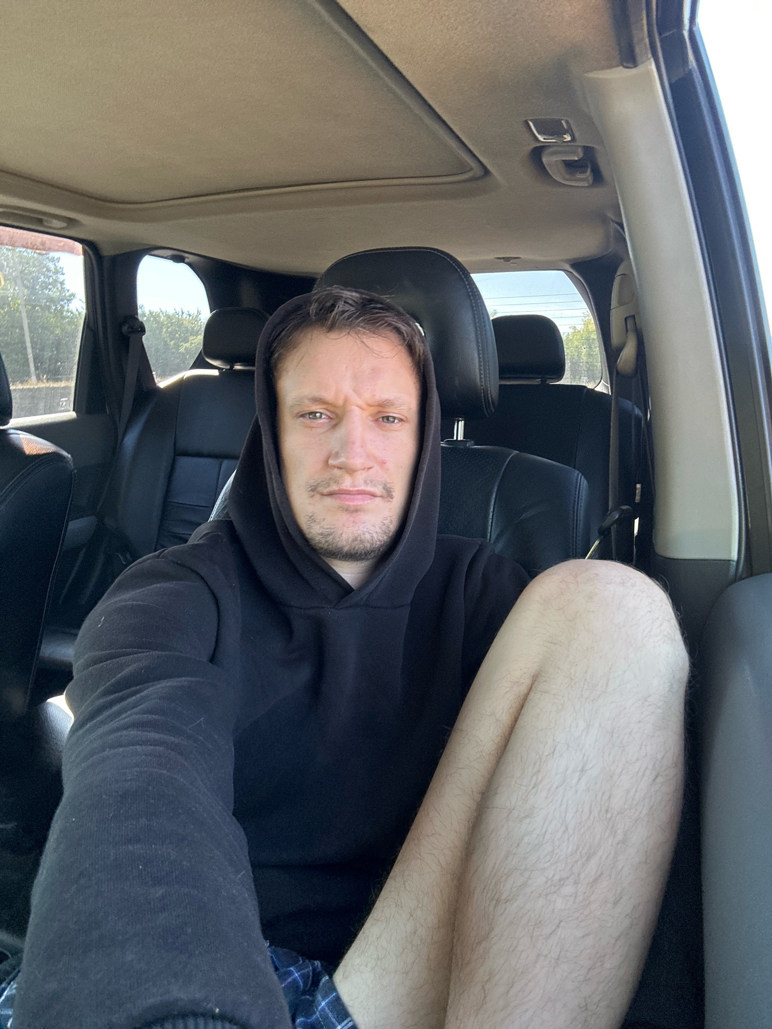 A man in a black hooded sweatshirt and checkered shorts sits in the passenger seat of a car. He is looking at the camera with a neutral expression. His left leg is crossed over his right leg. The interior of the car is visible, including the black leather seats and a sunroof. Outside the car, trees and a bright sky can be seen. The image appears to be taken in Makiv, Ukraine.