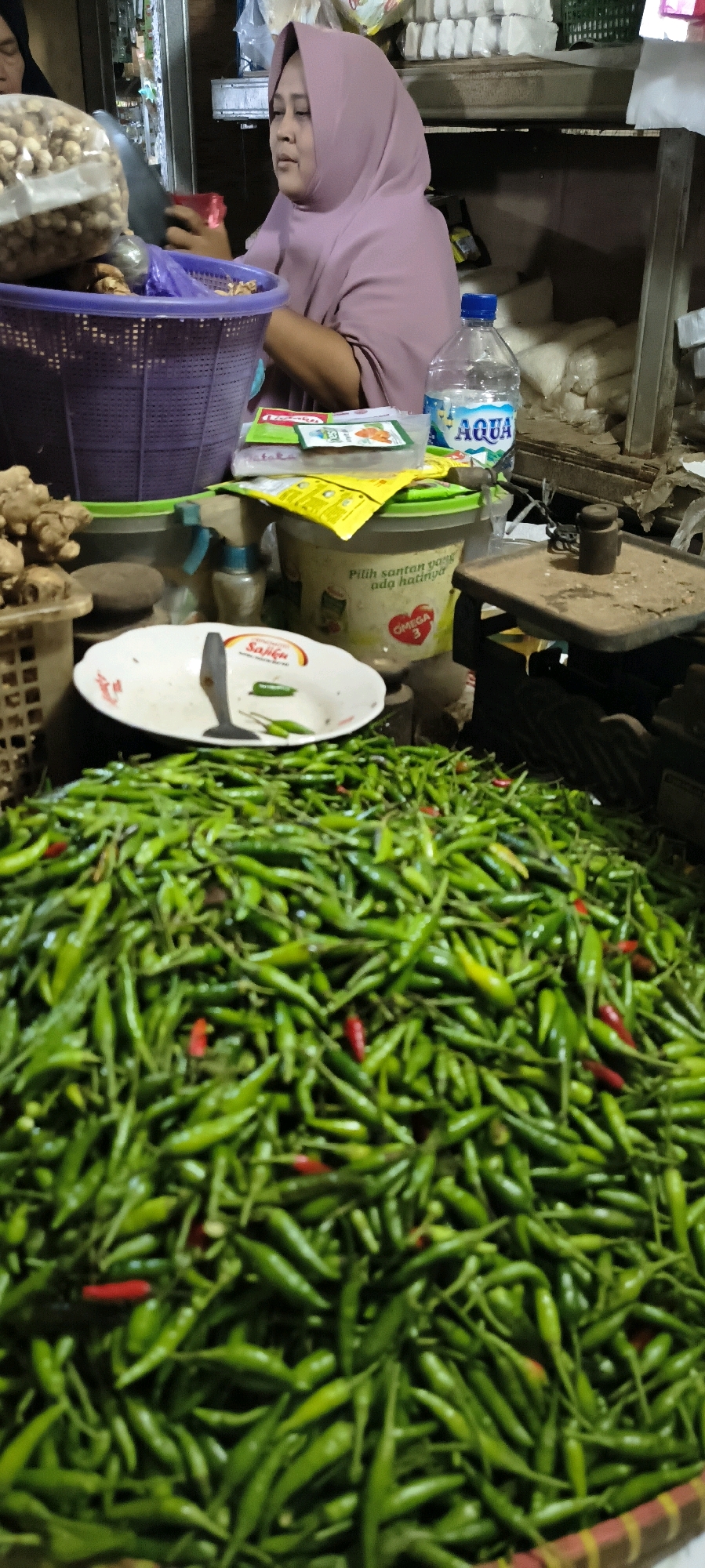 A woman wearing a lavender hijab is at a market stall in Weru Lor, Indonesia. She is looking down and appears to be engaged in a transaction, possibly weighing items on a scale behind her. In the foreground, a large pile of green chilies fills a basket. To the left of the chilies is a white plate with a metal utensil and a few more chilies. Behind the plate, a purple basket contains what appear to be nuts or seeds in a clear plastic bag. Various containers and packets of food items are visible on shelves in the background, including a bottle of Aqua water and packaging for cooking ingredients. The overall scene depicts a bustling marketplace environment.