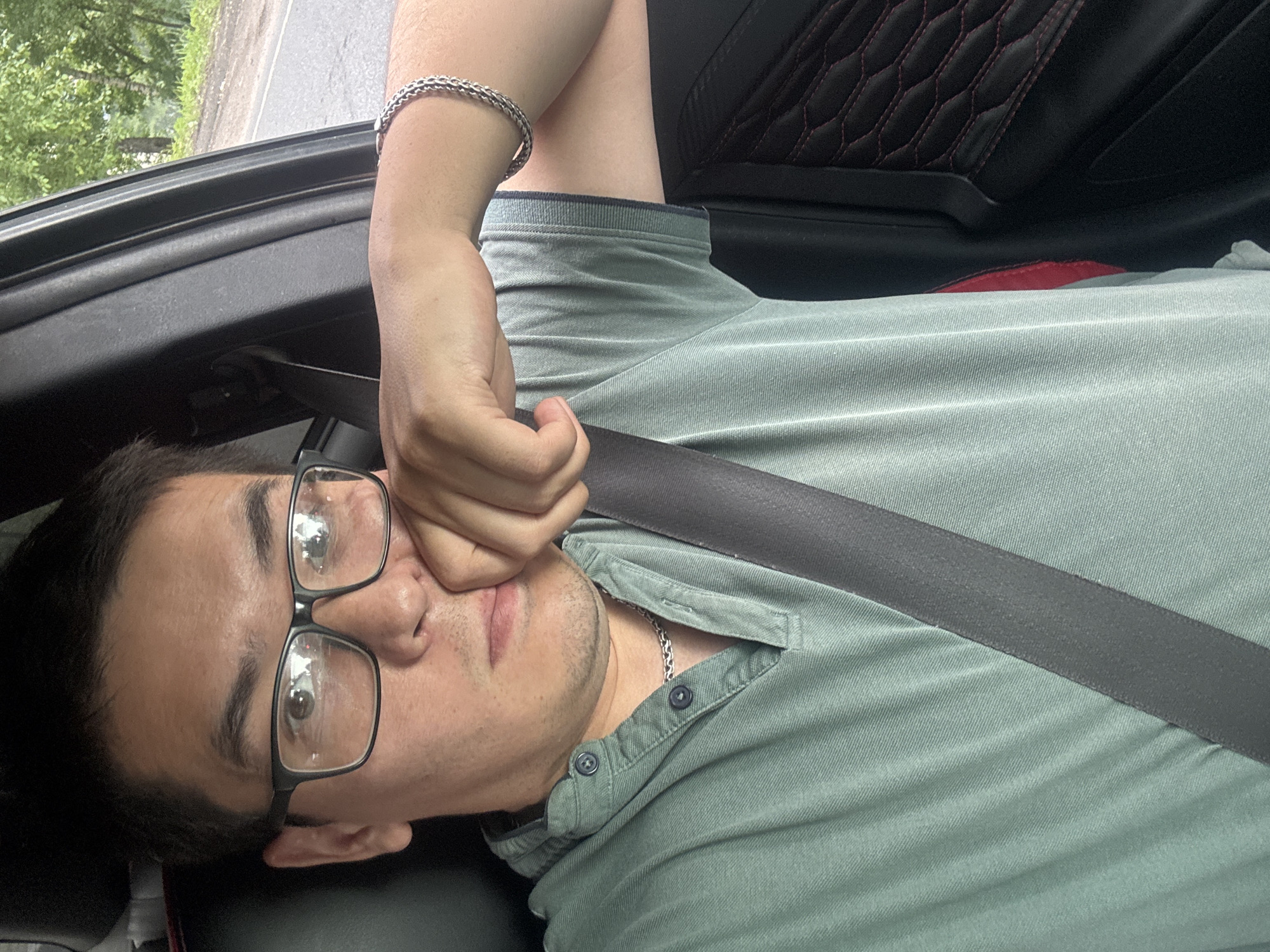 A medium shot captures a man sitting in the driver's seat of a car, with his right hand resting on his chin and his left arm on the car's door. He is wearing black-rimmed glasses, a light green collared shirt with two buttons, and a silver necklace. A seatbelt is visible across his chest. The car interior features black upholstery with red stitching on the door panel. Outside the car window, trees and a road can be seen, suggesting a rural or suburban environment. The lighting indicates it is daytime. The image was taken in Novoe Devyatkino, Russia.