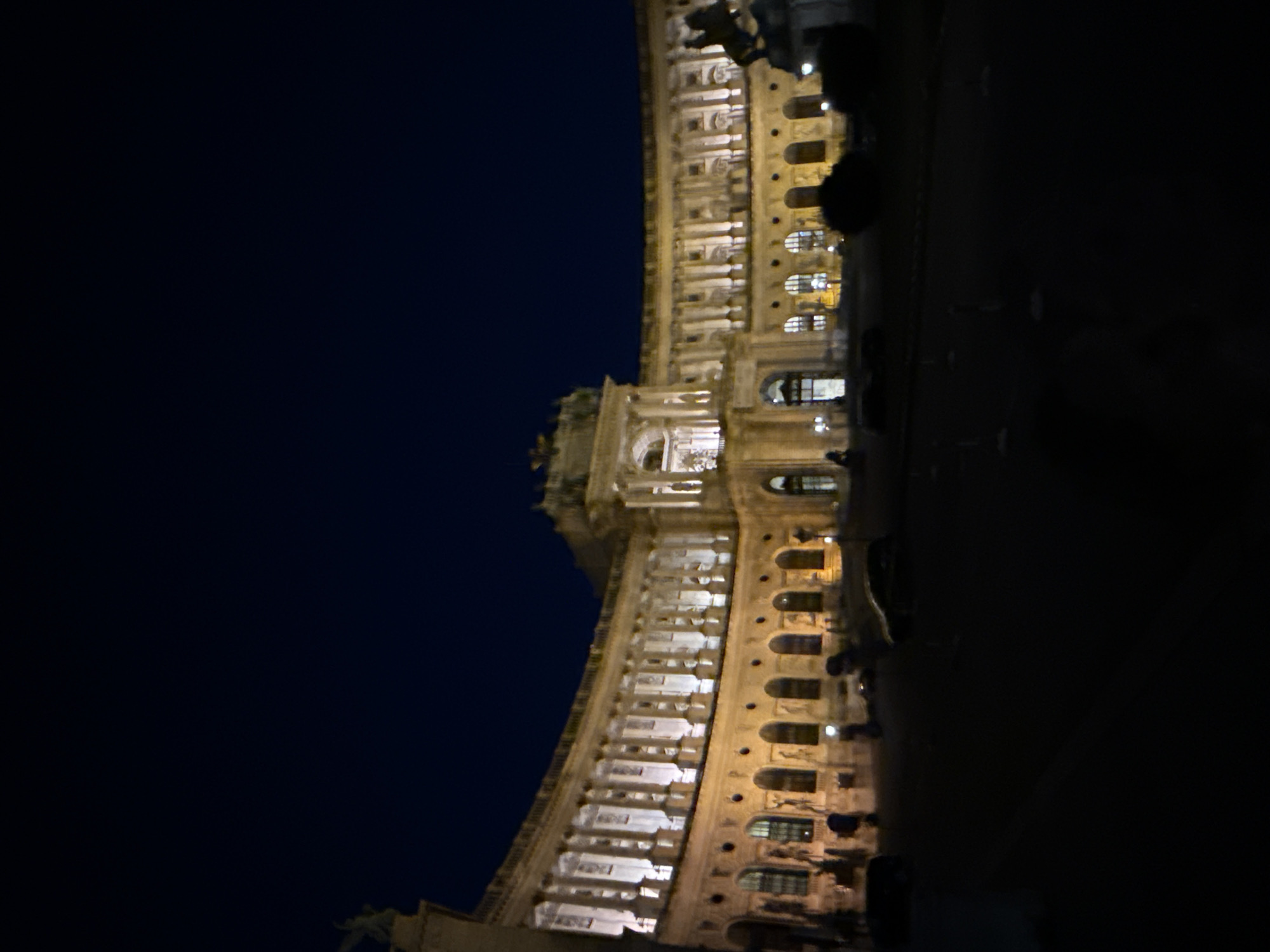 This is a nighttime photograph of the Hofburg Palace in Vienna, Austria. The grand Baroque building is illuminated by artificial lights, highlighting its arched windows, columns, and intricate architectural details. A dark, cloudless sky serves as the backdrop. In the foreground, the ground is dark and appears to be a paved area, possibly a plaza or courtyard. A few dark shapes on the ground could be vehicles, and there are some small, light-colored bollards or markers. The overall impression is one of grandeur and historical significance, amplified by the nighttime setting.