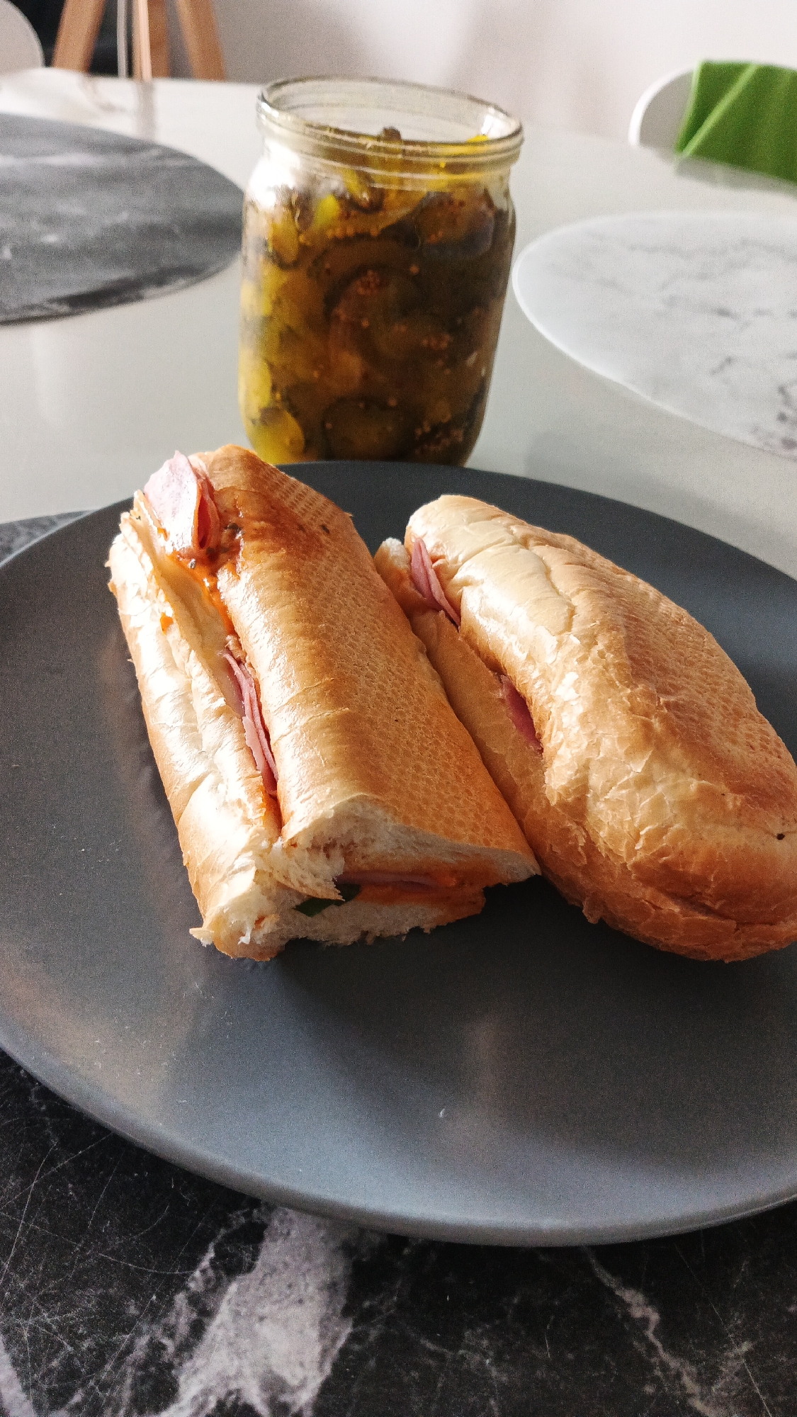 A close-up shot captures a dark gray plate with two halves of a submarine sandwich. The sandwich bread is golden brown and appears to be a baguette, sliced lengthwise and filled with pink meat, possibly ham, and a reddish sauce. In the background, a glass jar filled with sliced pickles and mustard seeds sits on a white surface, possibly a table. The table surface is textured with a black and white marble-like pattern. The lighting suggests an indoor setting. The scene does not provide any specific clues to its location in Zielona Góra, Poland.