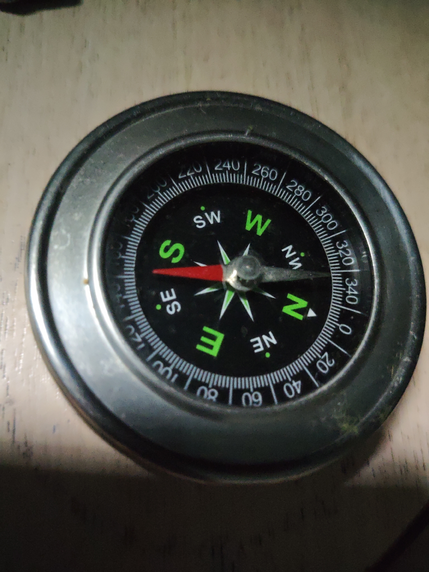 A close-up, overhead shot shows a black and silver compass with a metallic bezel, resting on a light-colored, textured surface, possibly wood. The compass face is marked with cardinal directions (N, S, E, W, NE, SE, SW, NW) and degree markings from 0 to 360. A red needle, indicating direction, points towards the 'S' marking. The image is slightly blurry and dark, with a narrow beam of light illuminating the center of the compass. There are no people, actions, or interactions visible in the image. The location of Naberezhnye Chelny, Russia is not determinable from this image.