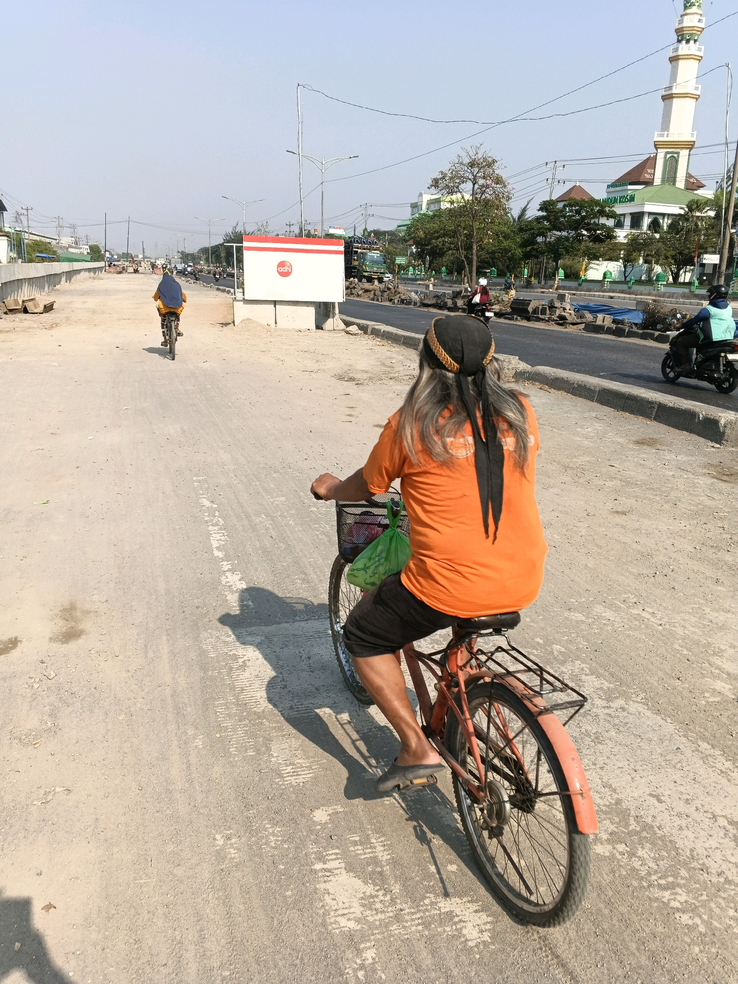 An elderly man with long grey hair, wearing an orange t-shirt, black shorts, and a black headscarf, cycles on a dirt road in Semarang, Indonesia. A green plastic bag is visible in the basket of his bicycle. In the background, a mosque with a white minaret and green dome stands prominently. Construction is evident along the roadside, with barriers and piles of dirt and materials. Other individuals on bicycles and motorcycles are also present, contributing to the busy urban scene. The weather appears to be clear and sunny.