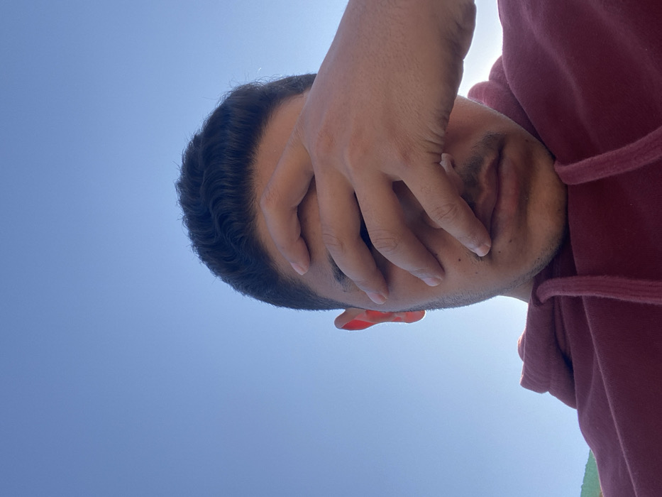 A man in a maroon hoodie shields his eyes from the sun. His hand covers much of his face, obscuring details except for part of his ear and mouth. He has dark hair with subtle waves. The background is a bright blue sky, indicating a sunny day. The perspective is from below, looking up at the man.