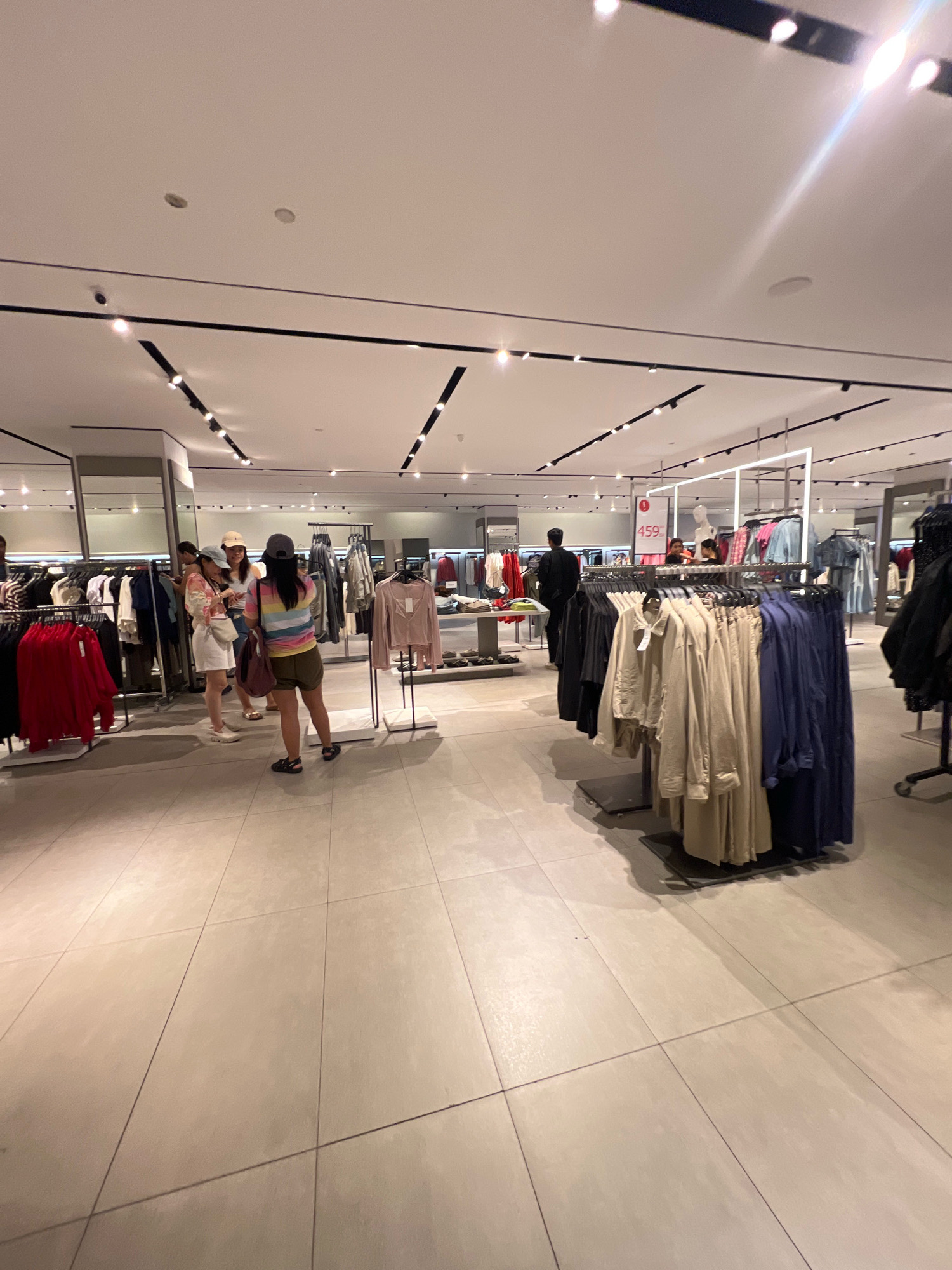 This is a wide-angle, eye-level shot of the interior of a clothing store in the city of Kuta, Indonesia. The store appears to be a contemporary retail space with bright, modern lighting and a polished tiled floor. Numerous racks of clothing are visible, arranged in sections, with a variety of garments hanging. In the foreground, to the left, a rack displays bright red garments. To the right, a rack holds a collection of beige and dark blue shirts or jackets. Several individuals are present in the store, some browsing clothing and others appearing to converse. In the mid-ground, a group of three women are standing together, seemingly examining items. Further back, a man in a black shirt is walking away from the camera. A price tag with "459" is visible on a display. The overall impression is of a busy, well-stocked retail environment.