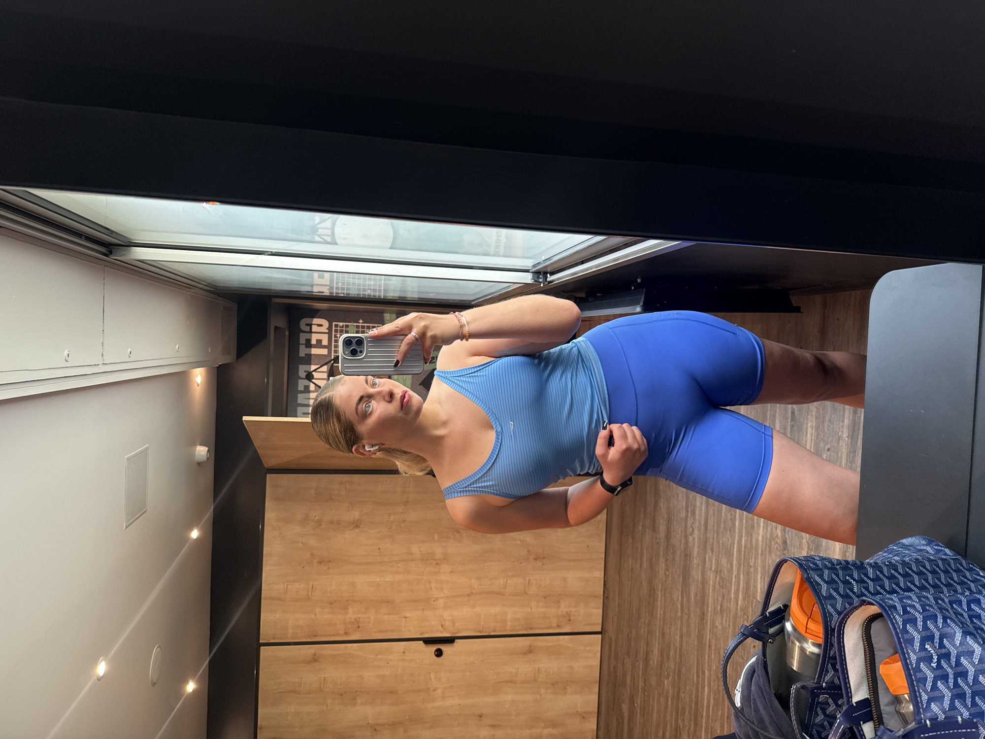 A young woman in workout attire takes a selfie in a gym locker room. She wears a blue ribbed tank top and matching blue biker shorts. Her blonde hair is pulled back. She holds an iPhone in her left hand and is looking at the screen. She has a bracelet on her left wrist and is wearing a black smart watch on her right wrist. In the background is a wall with built-in wooden lockers and overhead fluorescent lighting. A blue and white Goyard tote bag sits on the floor. The location is likely in Paris, France based on the visible lettering that contains "CEL" in reverse.