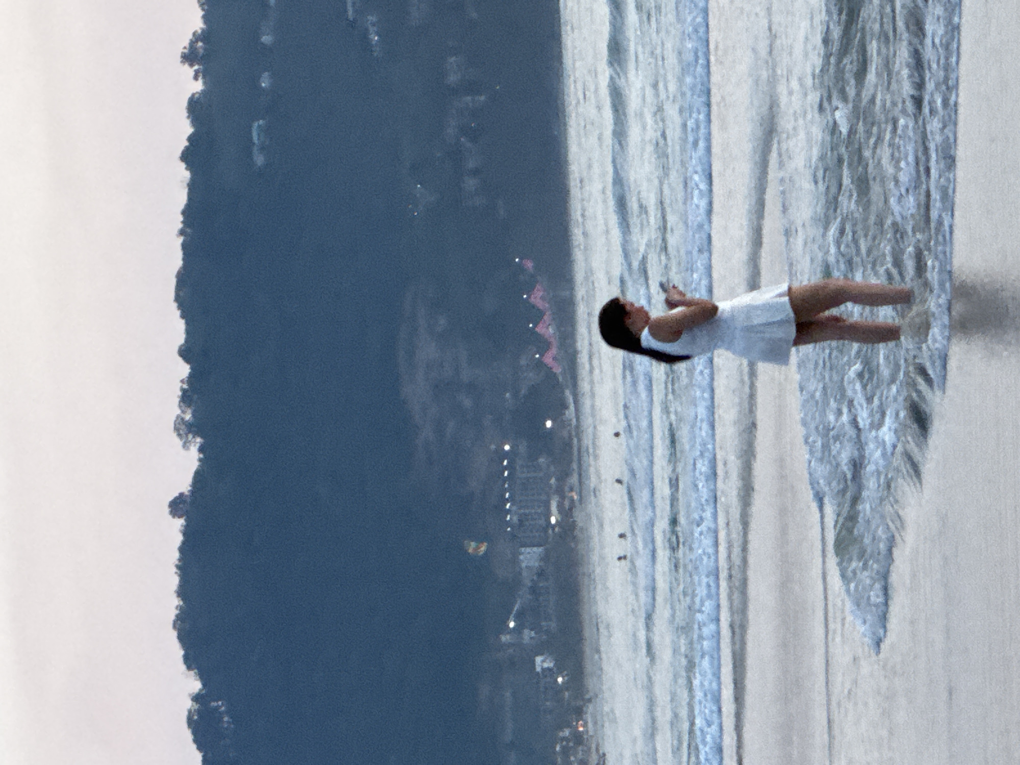 In Patong, Thailand, a young woman stands in the shallow water of the ocean, her white dress touching her knees. She is looking down at her phone, her dark hair falling over her shoulders. Gentle waves lap around her ankles, and further out, other people can be seen in the water. In the background, a tree-covered hill rises above a town with lights twinkling in the distance. The sky is a soft, hazy grey, suggesting either dawn or dusk.