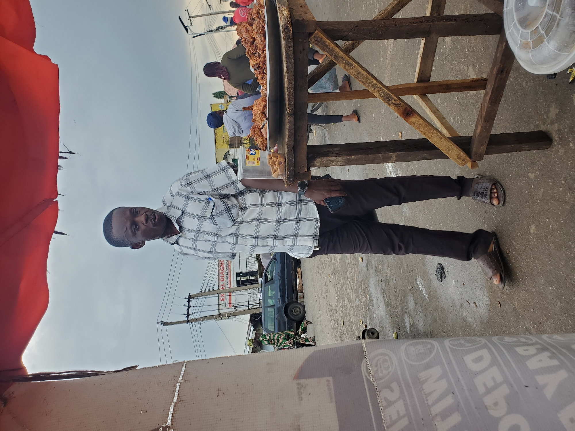 A man in a plaid shirt and dark pants stands in front of a food stall in Gwong, Nigeria. The food stall, made of rough-hewn wood, is laden with a large quantity of fried chicken. Other people are visible in the background, some attending the stall and others walking by. A blue van is parked on the side of the road. The scene appears to be outdoors during the day, with a bright, overcast sky. There are utility poles with wires visible in the distance. Some text is visible on a sign behind the food stall, reading "EXCEL DIAGNOSTIC CLINICAL SERVICE." Another sign with text is visible on a cardboard box in the foreground.