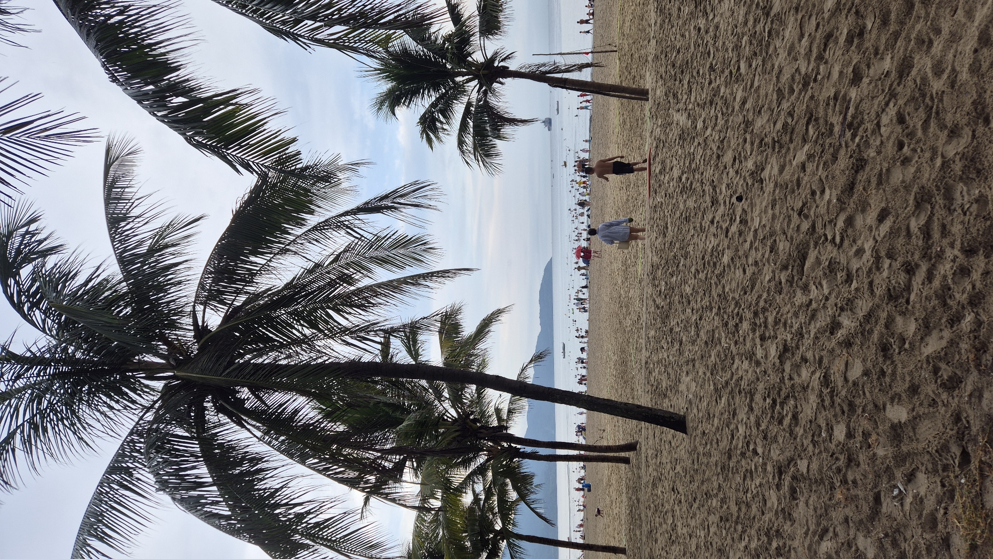 This image depicts a crowded beach scene at My Khe Beach in Đà Nẵng, Vietnam. The foreground is dominated by sand with visible footprints and a few scattered objects. In the mid-ground, a group of people are scattered across the beach, some walking, others possibly engaged in recreational activities. Palm trees with lush fronds are prominent, framing the scene and casting shadows on the sand. The ocean stretches out to the horizon, with several boats visible in the distance. The sky is overcast, suggesting a potentially hazy or cloudy day. The overall atmosphere is one of a busy public beach with numerous visitors enjoying the seaside.